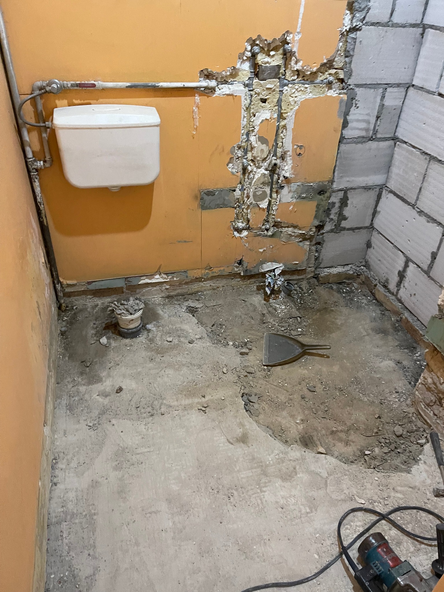 Łazienka w trakcie demontażu: odsłonięte rury, fragment ściany z cegieł, kuweta z narzędziami, szpachelka i brudna podłoga przed remontem.