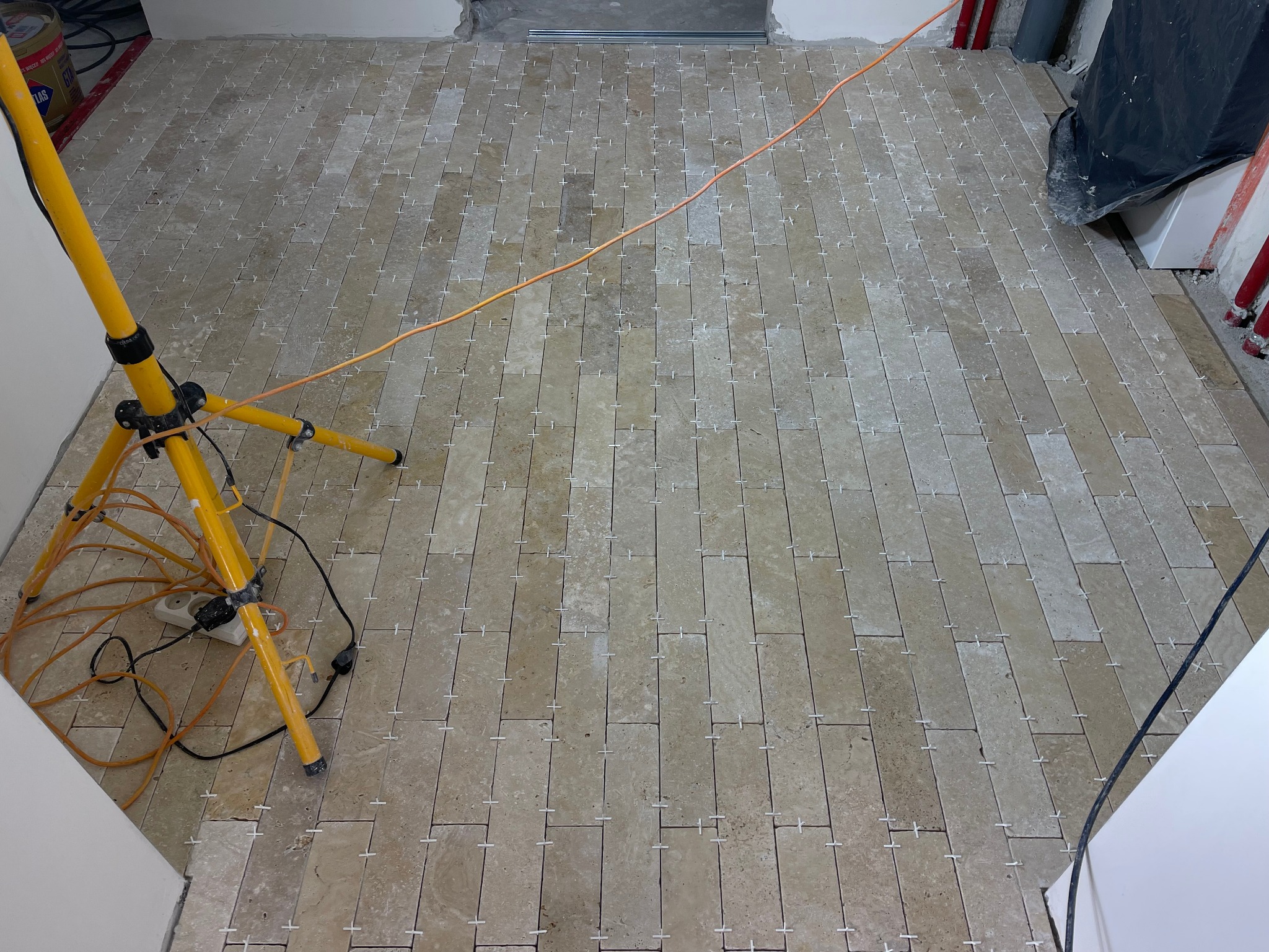 Ułożone płytki podłogowe w trakcie fugowania, widoczne krzyżyki dystansowe, oświetlenie budowlane na trójnogu i przewody elektryczne.