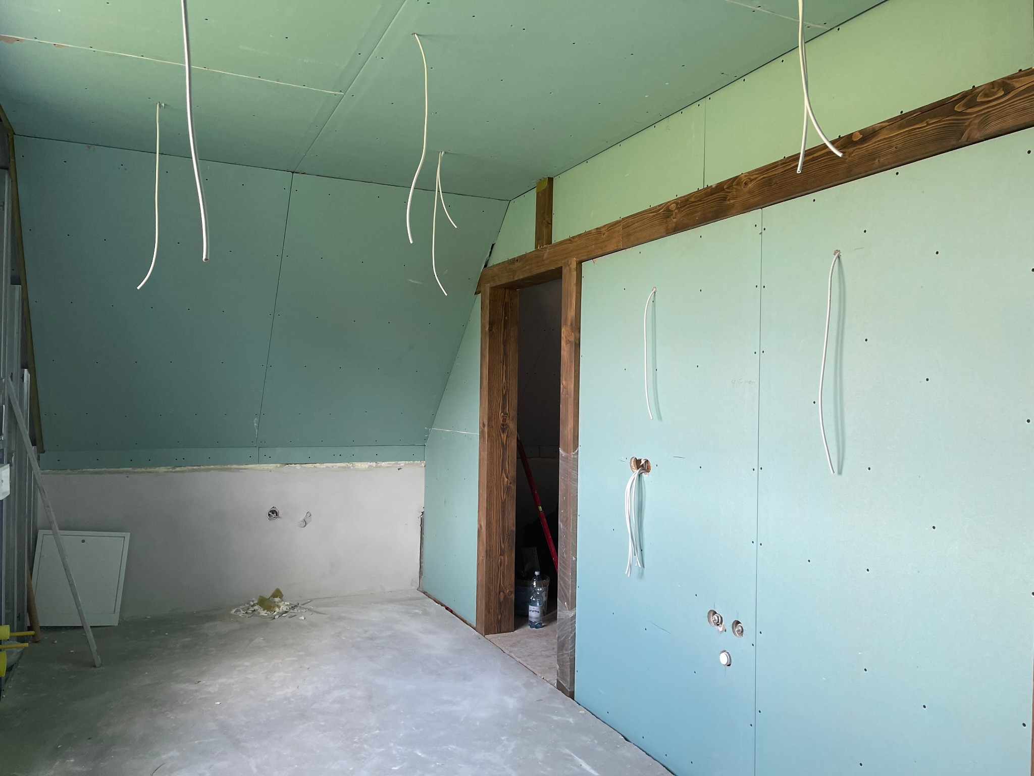 Poddasze w trakcie prac wykończeniowych: zamontowane płyty karton-gips, widoczne kable zwisające z sufitu, surowe drewniane ramy drzwiowe i fragment ściany przygotowanej do dalszych prac.