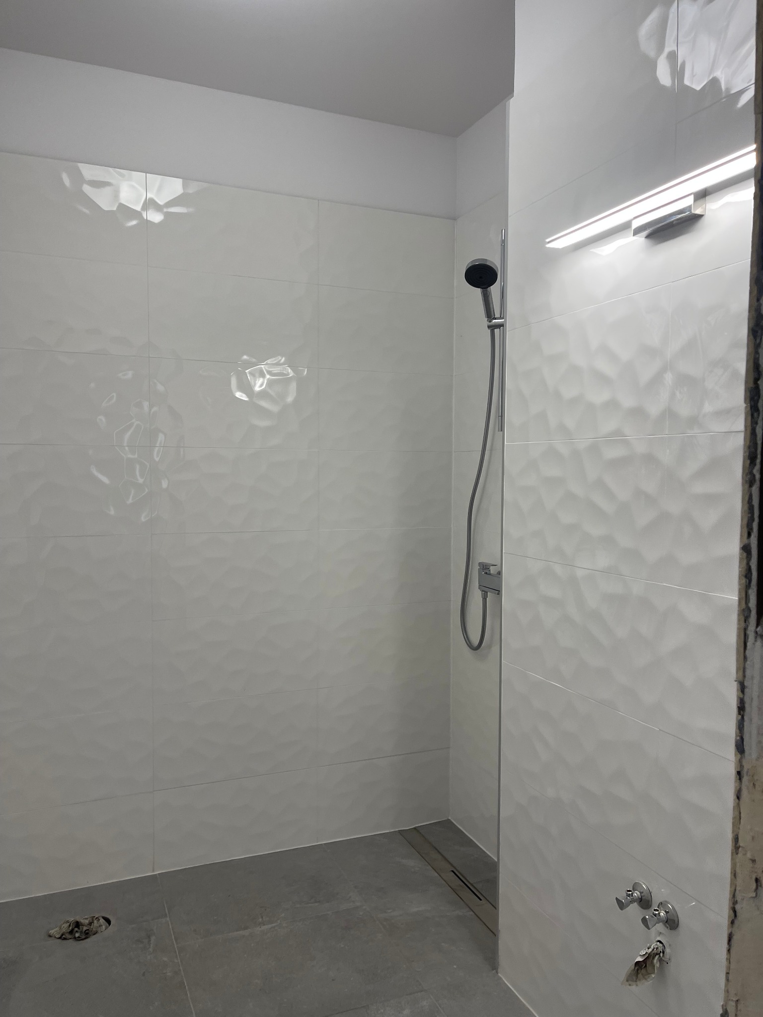 Nowoczesna kabina prysznicowa z białymi, trójwymiarowymi płytkami, szarymi płytkami podłogowymi z odpływem liniowym i chromowaną armaturą, oświetlona listwą LED, w trakcie remontu.