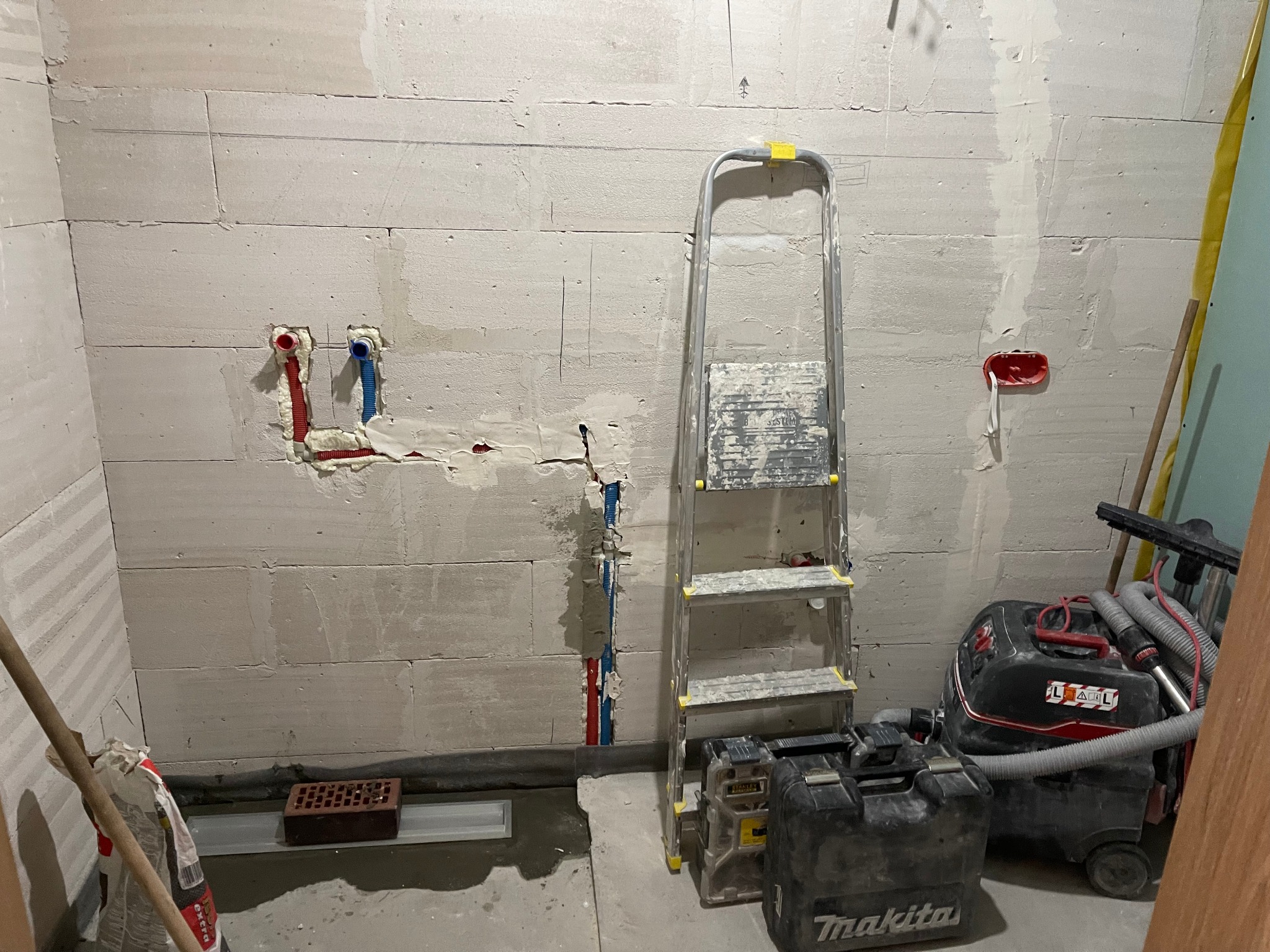 Ściana z betonu komórkowego w trakcie przygotowywania do wykończenia, widoczne instalacje wodne z rurami w kolorze czerwonym i niebieskim, drabina aluminiowa, odkurzacz przemysłowy i skrzynki...