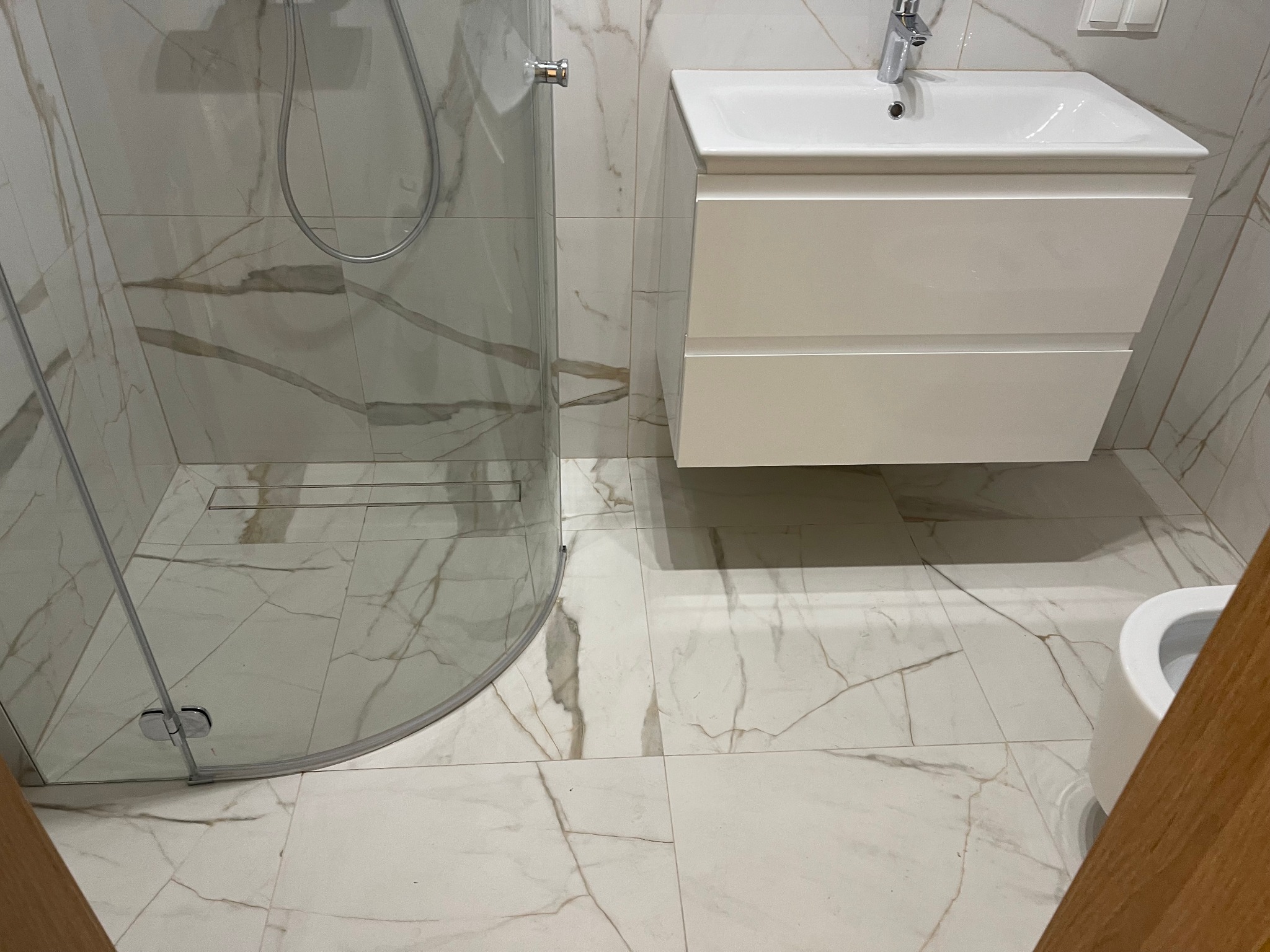 Nowoczesna łazienka z kabiną prysznicową typu walk-in, białą szafką pod umywalką i płytkami imitującymi marmur z wyraźnym żyłowaniem.