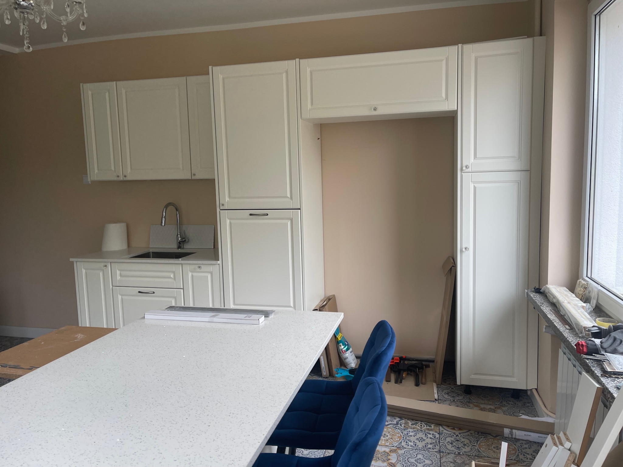 Montaż białych szafek kuchennych z blatem o wzorze lastryko, widoczne narzędzia i elementy wykończeniowe na podłodze z dekoracyjnymi płytkami.