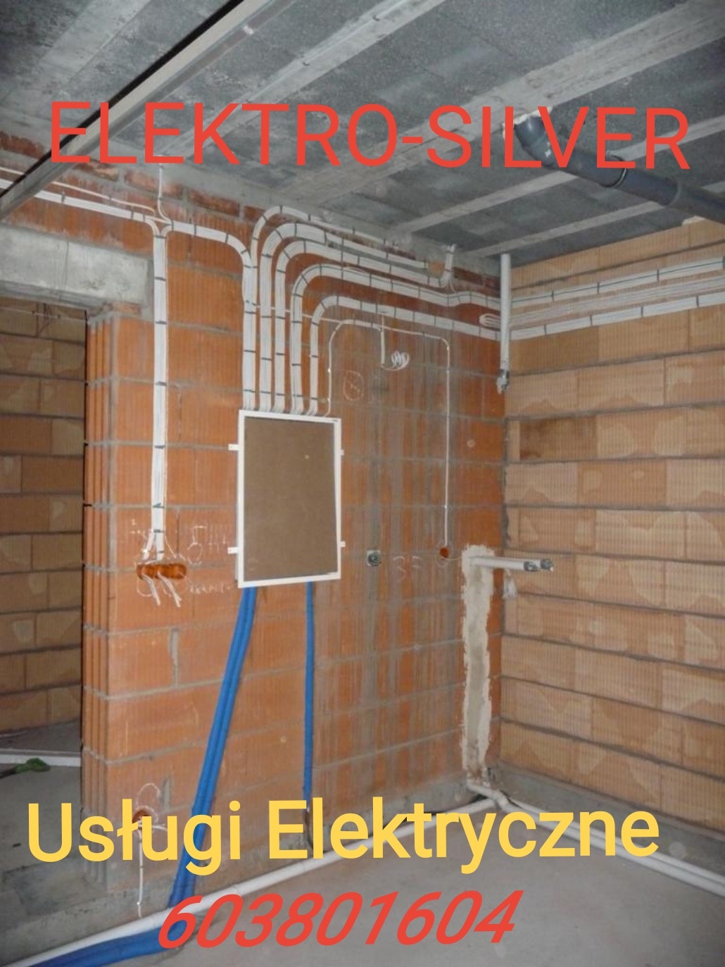 Wnętrze w stanie surowym z widoczną instalacją elektryczną: kable w białych rurkach prowadzone po ścianach z cegły, skrzynka rozdzielcza z brązowymi drzwiczkami, niebieskie rury PCV przy podłodze...