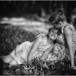 M.W.FotoPasja - Czarno-białe zdjęcie uśmiechniętej dziewczynki leżącej na trawie, obejmowanej przez kobietę, która całuje ją w głowę, wykonane w plenerze.