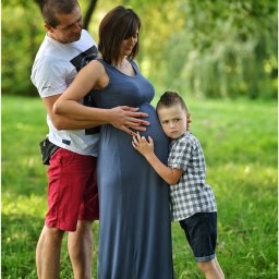 M.W.FotoPasja - Przyszła mama w długiej szarej sukience, obejmowana przez męża i syna na tle zielonego parku.