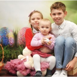 M.W.FotoPasja - Trójka uśmiechniętych dzieci pozuje na tle wielkanocnej dekoracji z koszykiem pełnym jajek i pluszowym króliczkiem z różową spódniczką.
