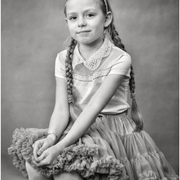 M.W.FotoPasja - Czarno-białe portretowe ujęcie uśmiechniętej dziewczynki z warkoczami, siedzącej na krześle, ubrana w tiulową spódniczkę i bluzkę z koronkowym kołnierzykiem na neutralnym tle.