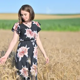 Młoda kobieta w czarnej sukience w kwiaty spaceruje po polu pszenicy, dotykając kłosów dłonią, z polami w tle.