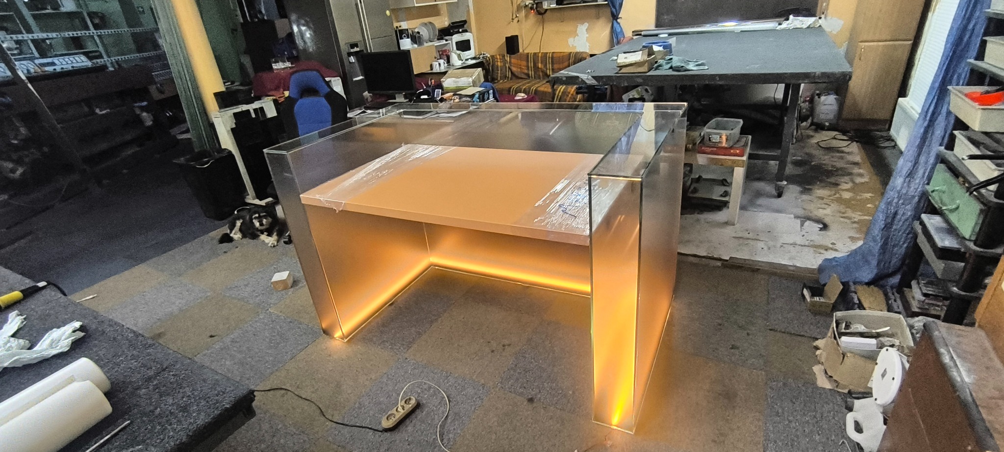 Szklany stół z podświetleniem LED w warsztacie, na blacie folia ochronna, obok leży pies i przedłużacz.