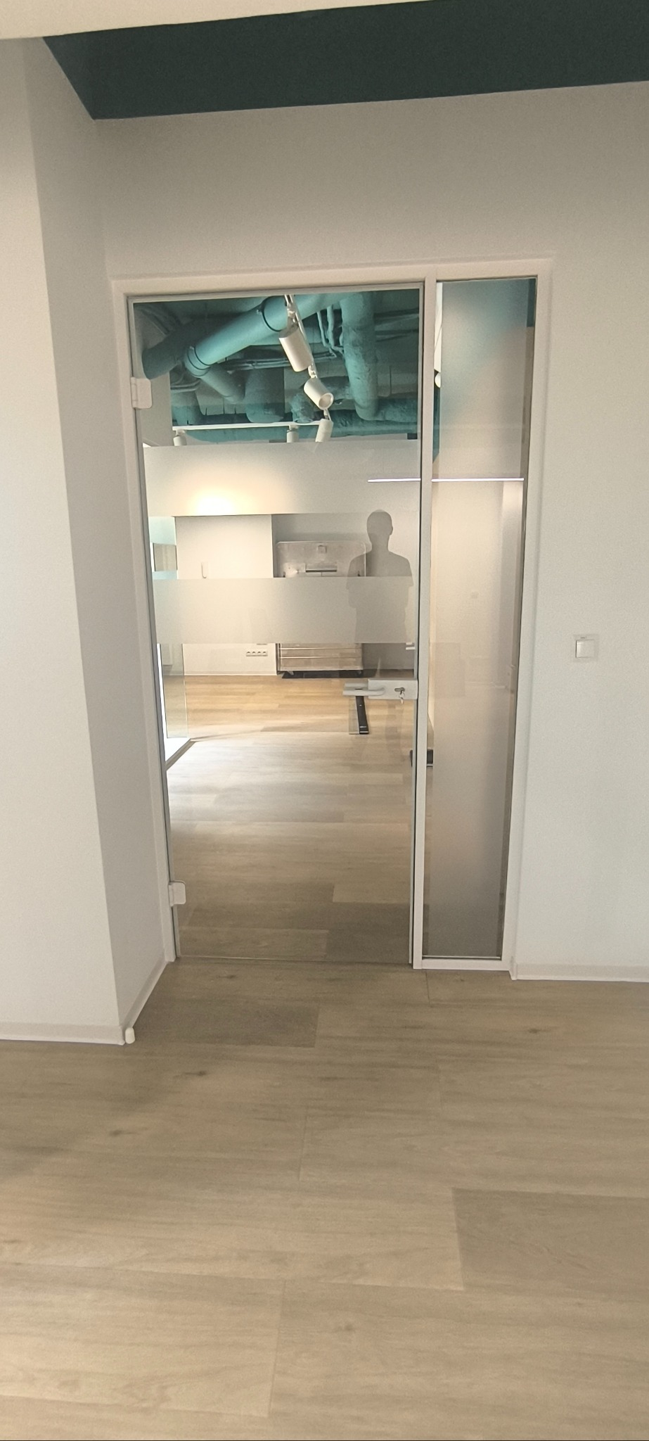 Szklane drzwi z matową folią w nowoczesnym biurze, widoczne rury pod sufitem i sylwetka osoby w tle.