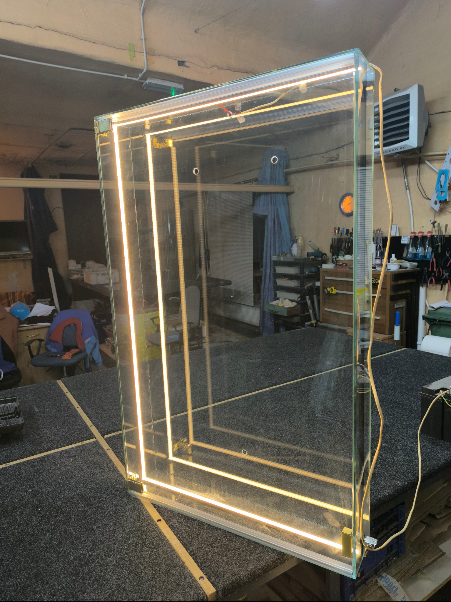 Szklana tafla z wbudowanym oświetleniem LED, ustawiona w warsztacie, widoczne przewody zasilające i odbicie wyposażenia warsztatowego w szkle.