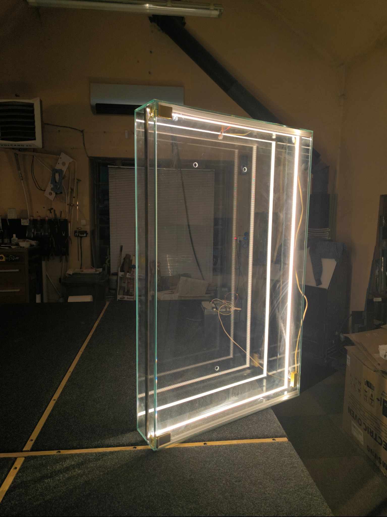 Szklana konstrukcja z oświetleniem LED w warsztacie, widoczne elementy montażowe i okablowanie, tło stanowią narzędzia i materiały.