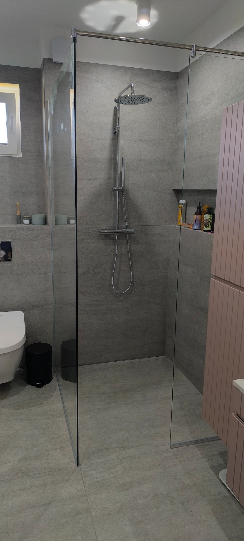 Nowoczesna kabina prysznicowa ze szklanymi ścianami i deszczownicą w łazience wykończonej w szarościach, z widoczną toaletą i półką z kosmetykami.