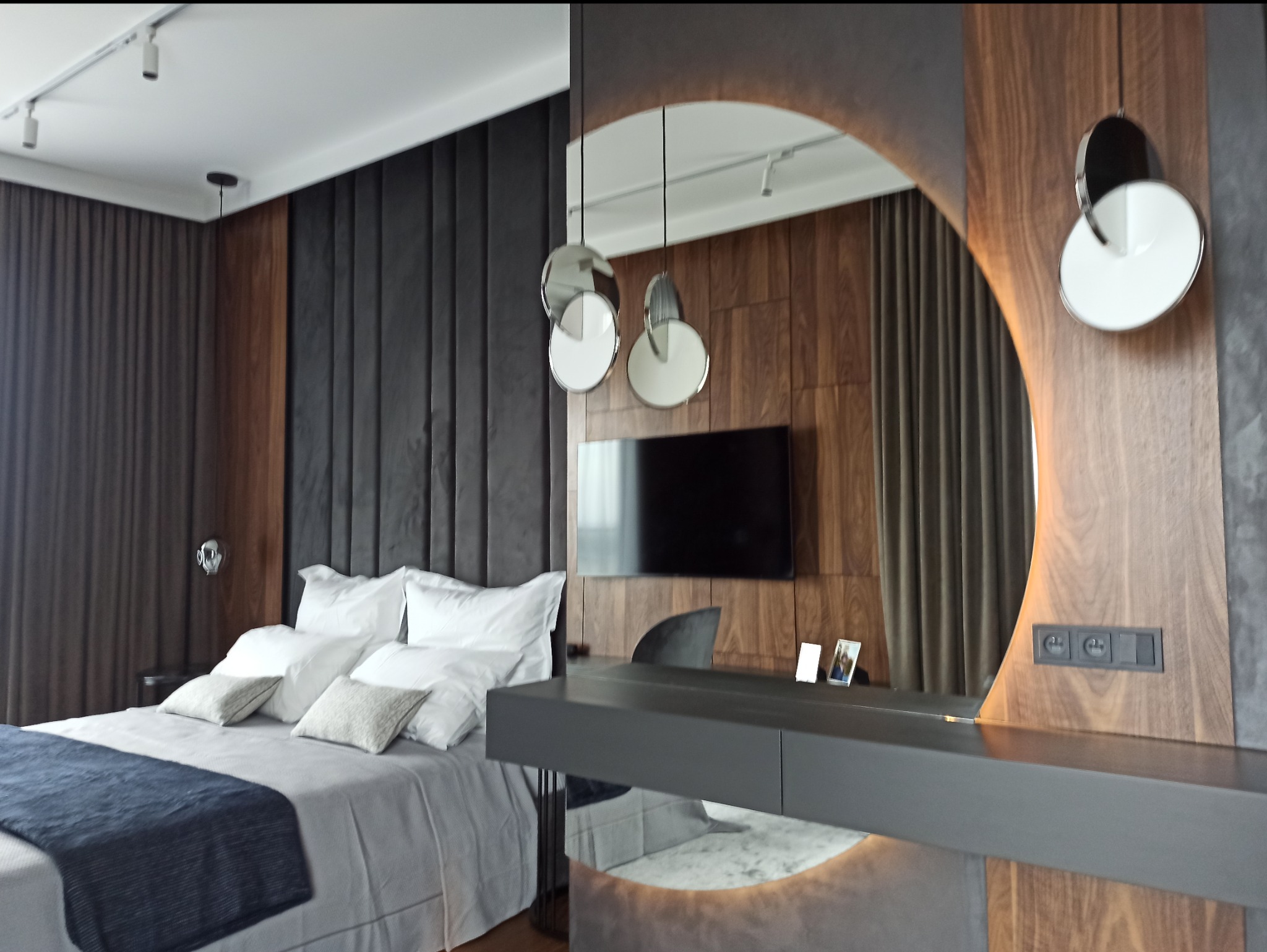 Nowoczesna sypialnia z łóżkiem, telewizorem i toaletką z owalnym lustrem z podświetleniem LED, wiszącymi lampami i drewnianą okładziną ściany.