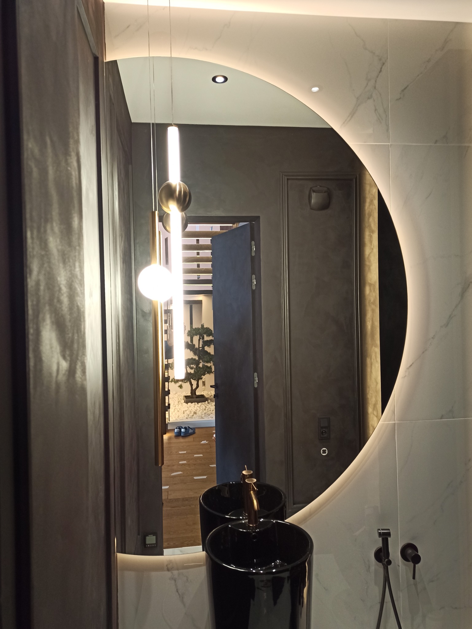 Okrągłe lustro z podświetleniem LED w łazience, odbijające wnętrze z dekoracyjnym drzewkiem bonsai i parą niebieskich butów na podłodze.