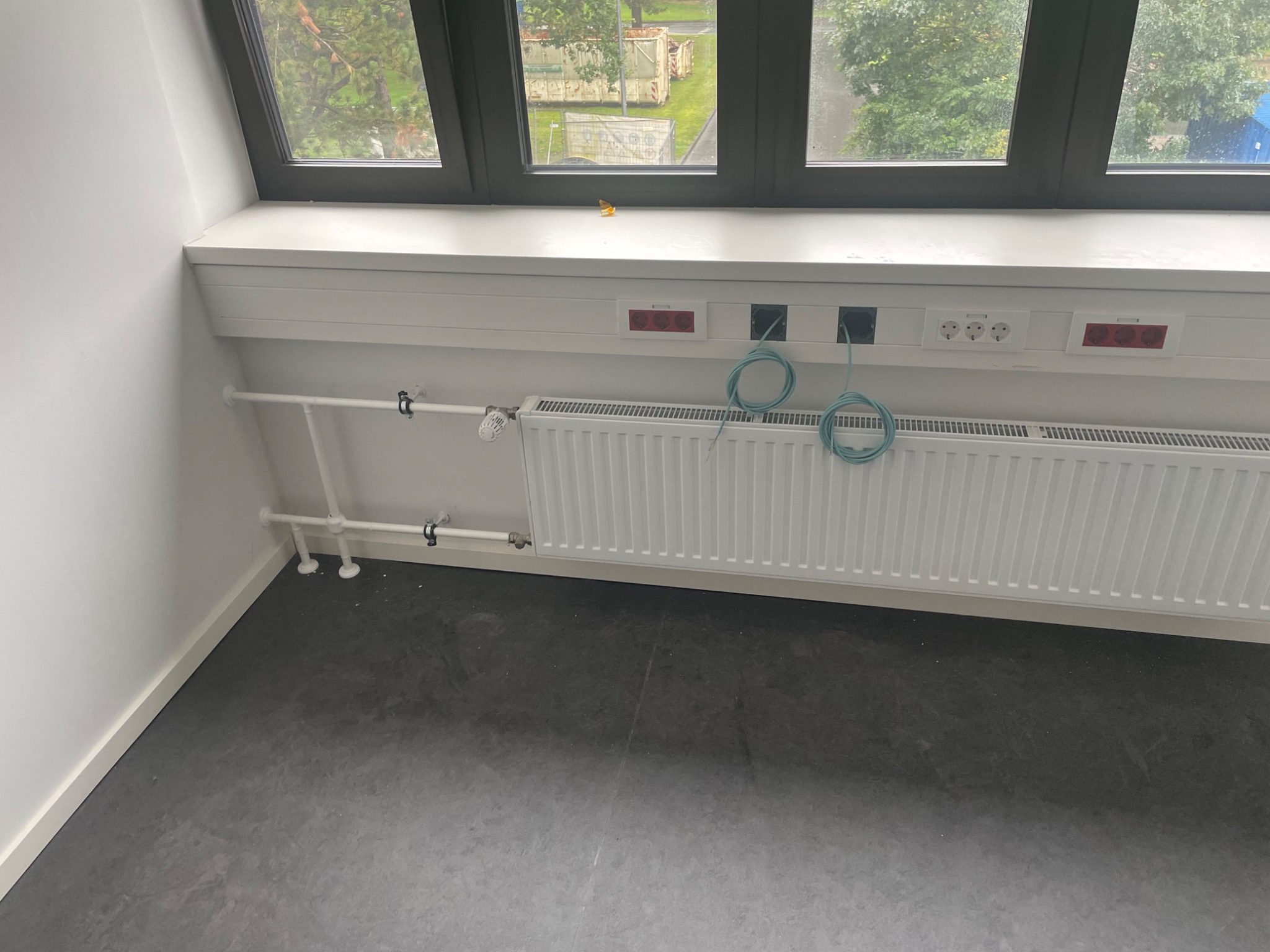 Biały grzejnik panelowy zamontowany pod oknem na poddaszu, z widocznymi rurami doprowadzającymi, gniazdkami elektrycznymi i kablami w kolorze turkusowym.