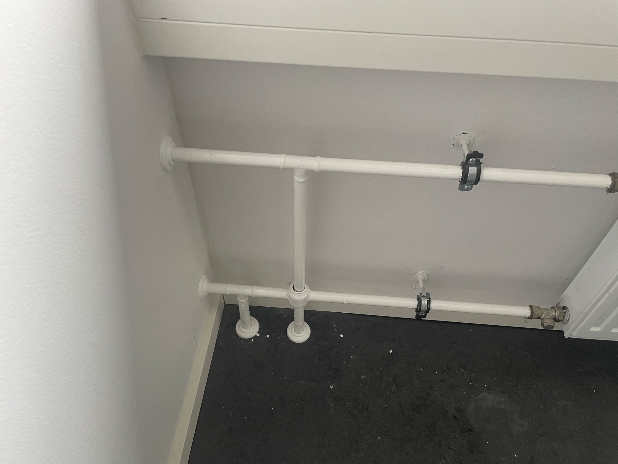 Białe rury grzewcze przymocowane do ściany i sufitu, widoczne elementy mocujące i fragment grzejnika, czarna podłoga.