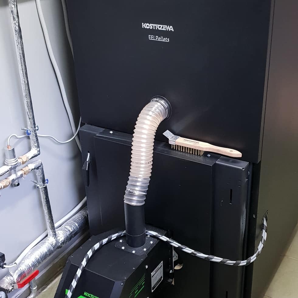 Czarny piec na pellet marki Kostrzewa z podłączonym przewodem i szczotką do czyszczenia na obudowie.