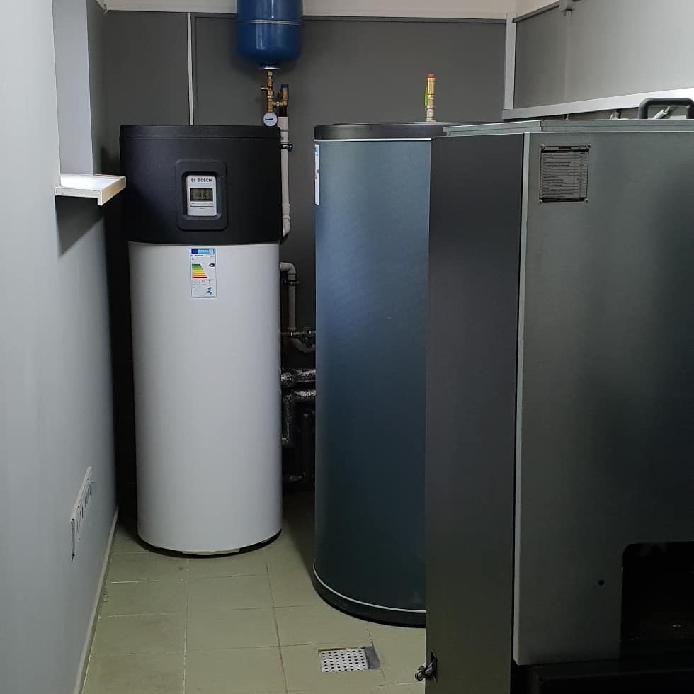 Biały zbiornik z czarnym panelem sterowania marki Bosch, obok szary zbiornik, niebieski zbiornik wyrównawczy na ścianie, instalacja w pomieszczeniu technicznym z szarymi ścianami i płytkami...