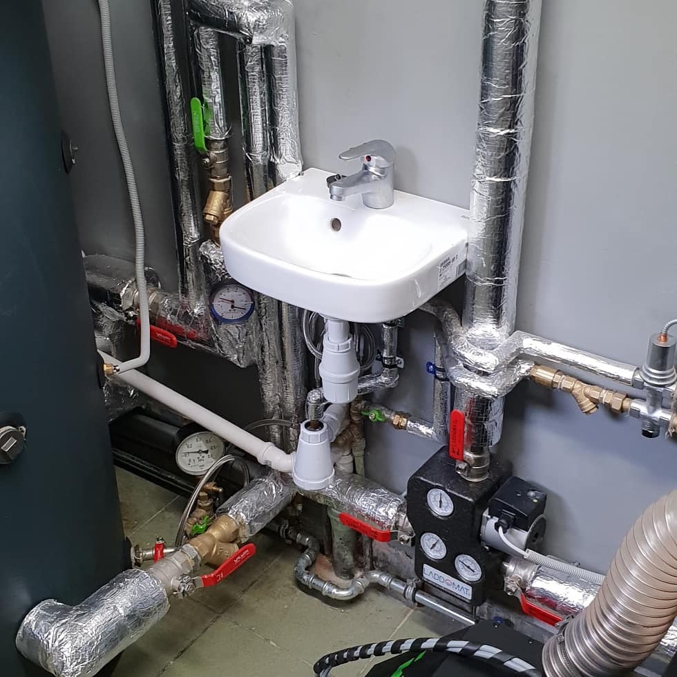 Niewielka biała umywalka zamontowana w kotłowni, otoczona rurami centralnego ogrzewania z izolacją, widoczny zasobnik wody i Laddomat z manometrami.