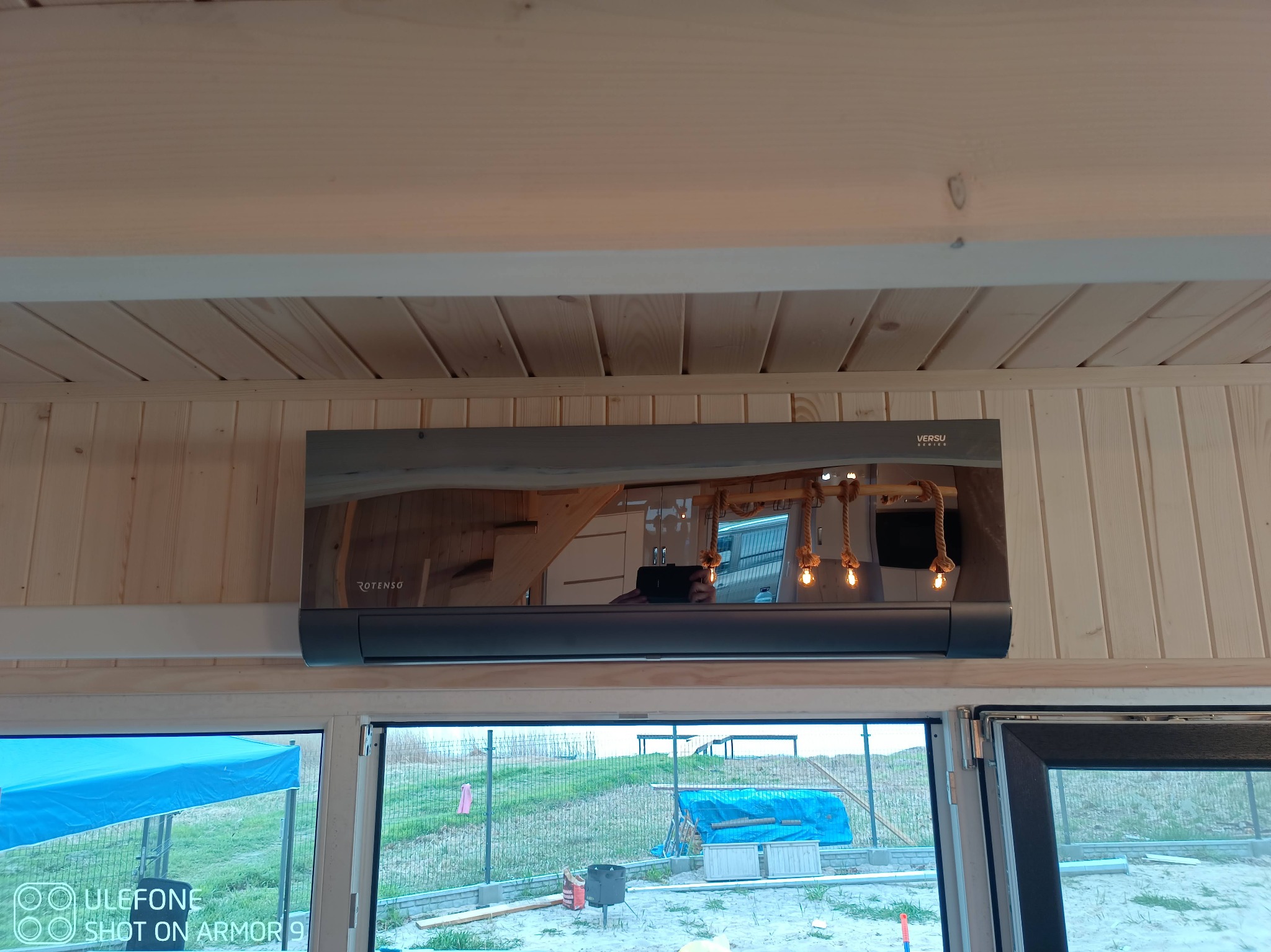 Szara jednostka wewnętrzna klimatyzatora Rotenso Versu zamontowana na drewnianej ścianie domku, widoczna przez okno zewnętrzna sceneria z niebieską plandeką i ogrodzeniem.