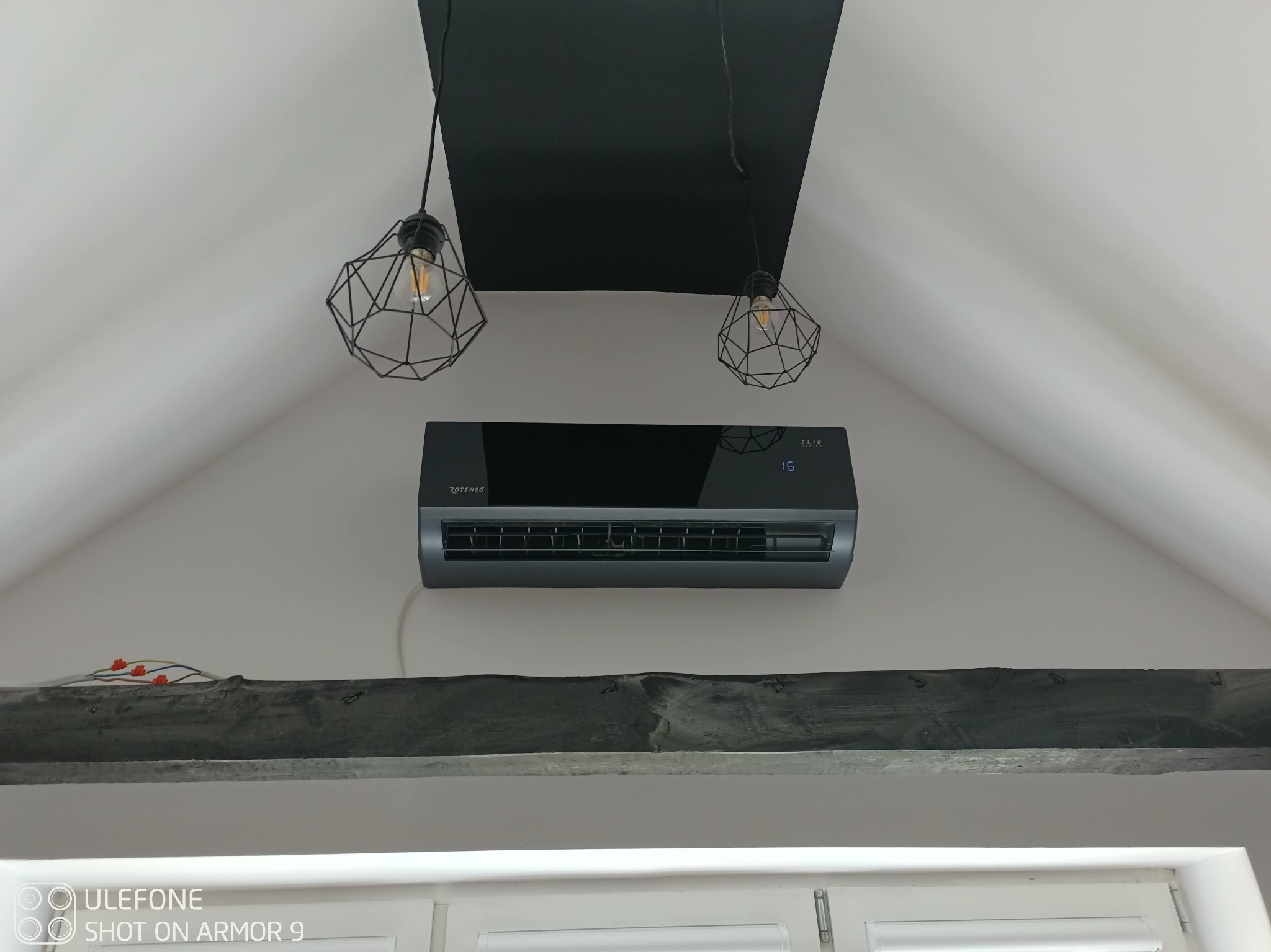 Czarny klimatyzator Rotenso zamontowany na ścianie pod skośnym sufitem w pokoju z drewnianą belką i nowoczesnymi lampami wiszącymi.