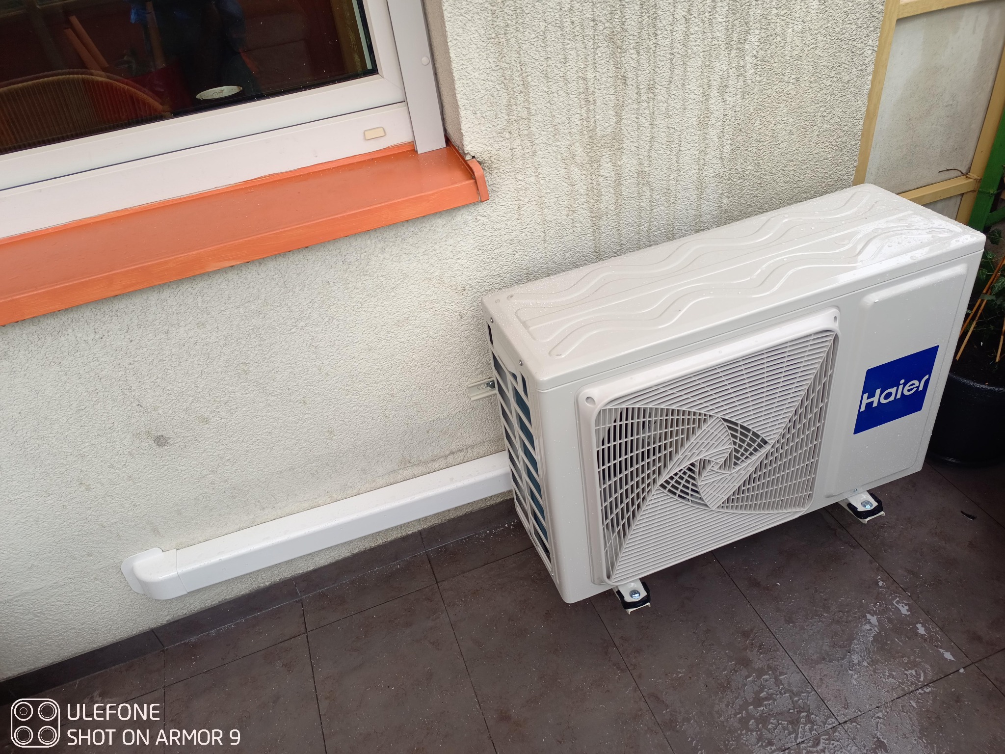 Zewnętrzna jednostka klimatyzacji Haier zamontowana na brązowej podłodze przy ścianie budynku z pomarańczowym parapetem okiennym.