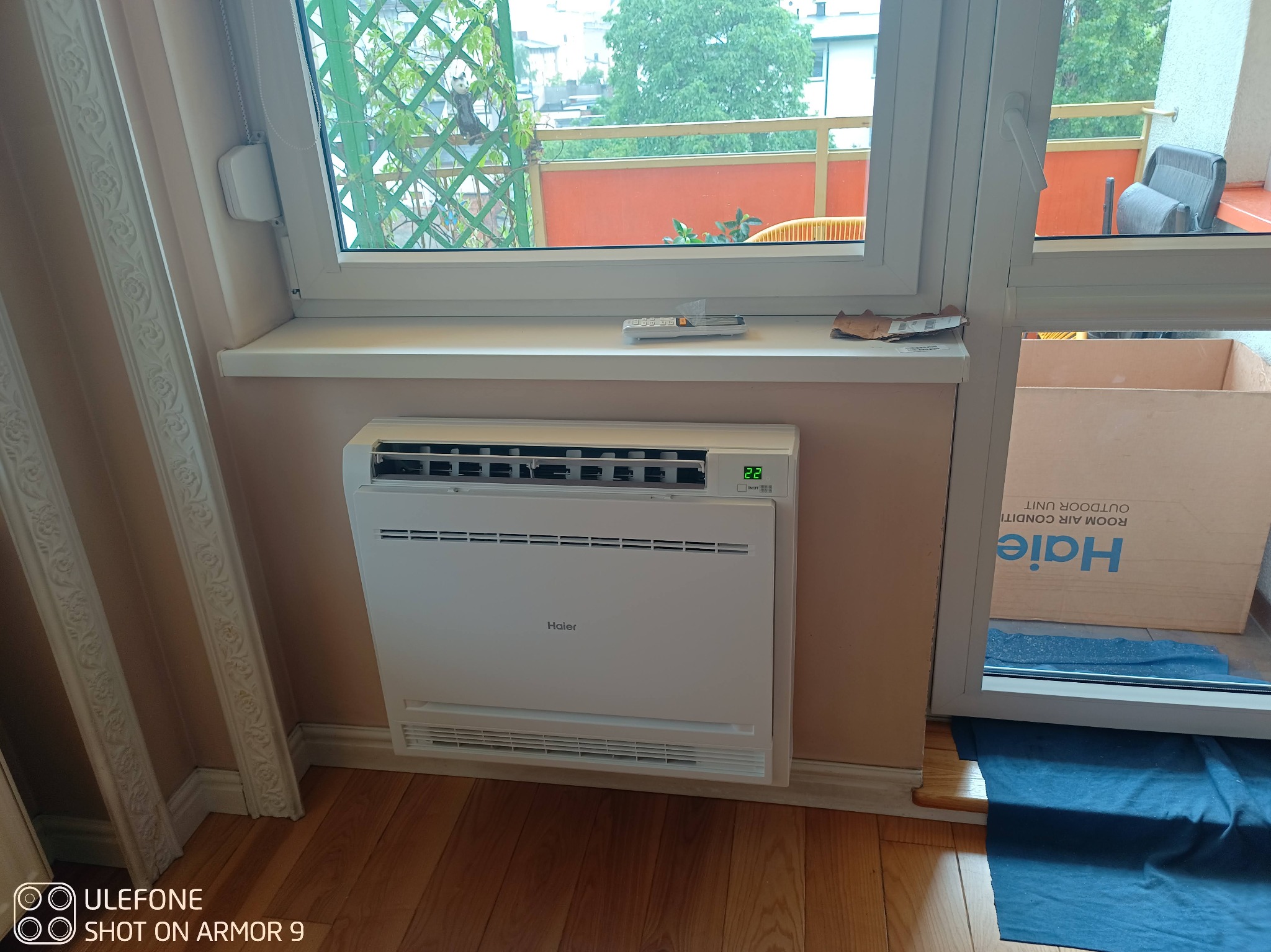 Biała, przypodłogowa jednostka klimatyzacyjna Haier zamontowana pod oknem z widokiem na balkon, pilot na parapecie, częściowo widoczne kartonowe opakowanie jednostki zewnętrznej.