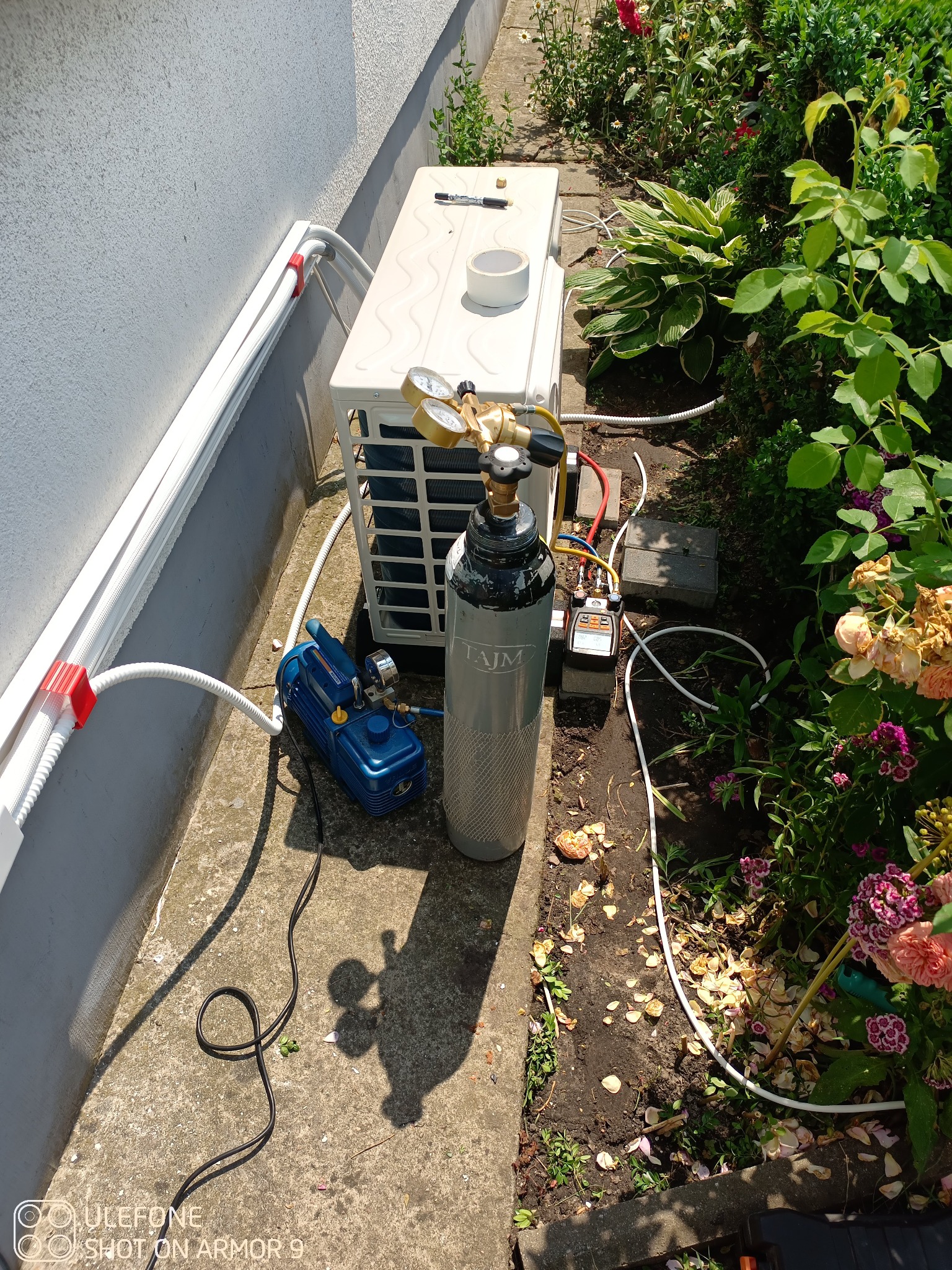 Zewnętrzna jednostka klimatyzacji podczas podłączania, widoczna butla z gazem, manometry i pompa próżniowa, tło z roślinnością ogrodową.