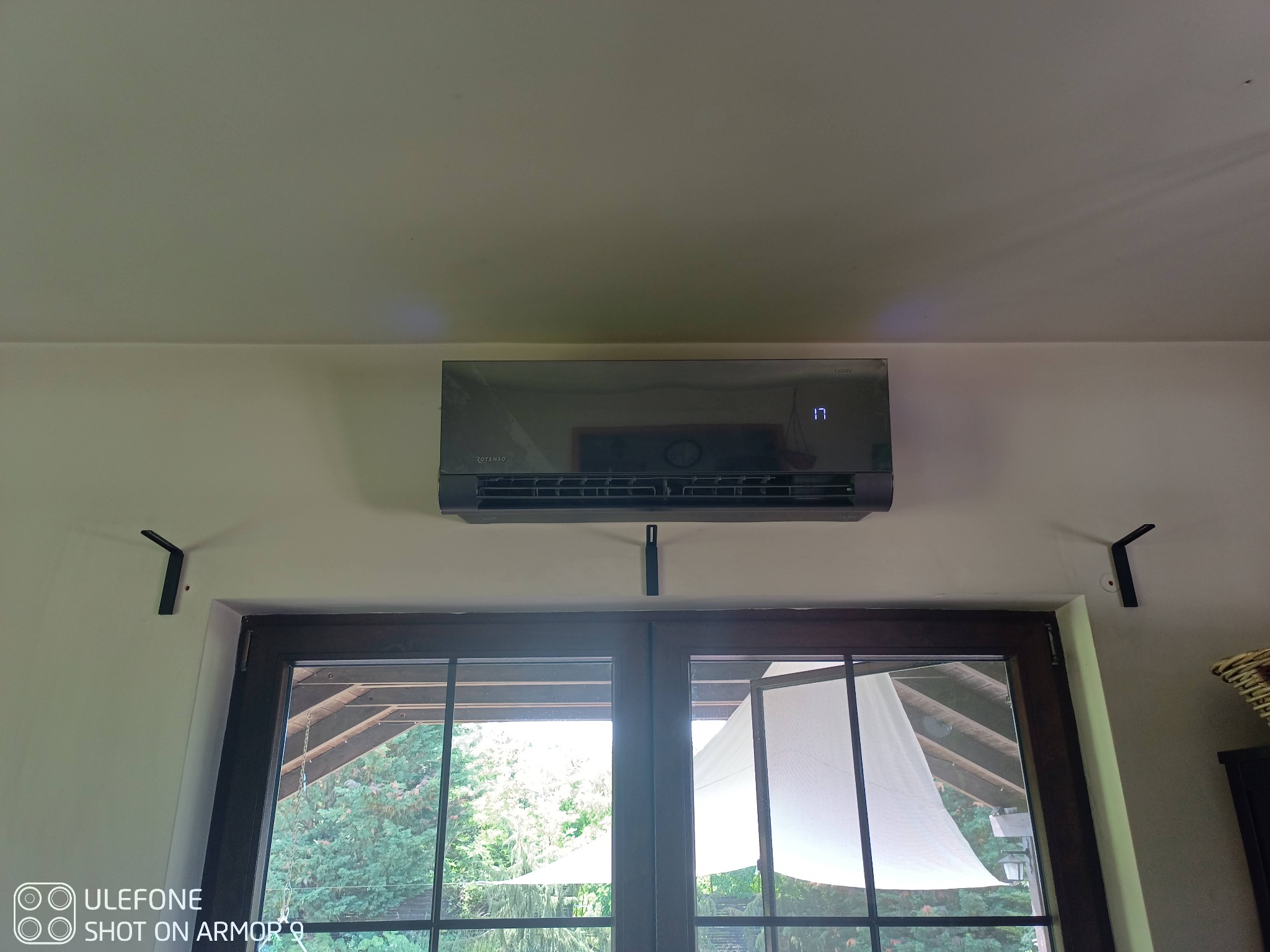 Czarny klimatyzator ścienny z wyświetlaną temperaturą 17 stopni, zamontowany nad oknem z widokiem na zieleń.