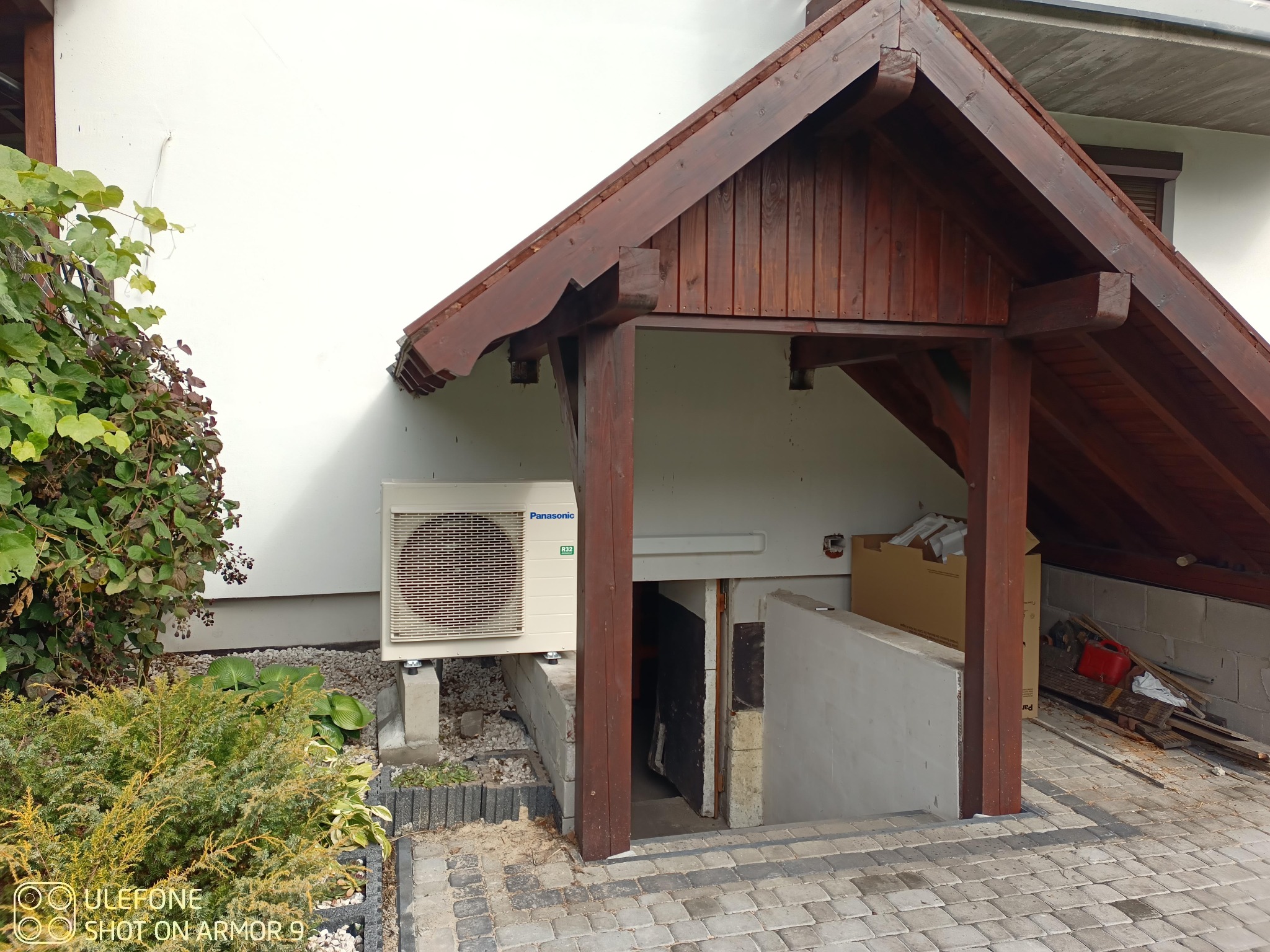 Zewnętrzna jednostka klimatyzacji Panasonic zamontowana na betonowych podstawach przy wejściu do piwnicy z drewnianym zadaszeniem, widoczne rośliny i kostka brukowa.