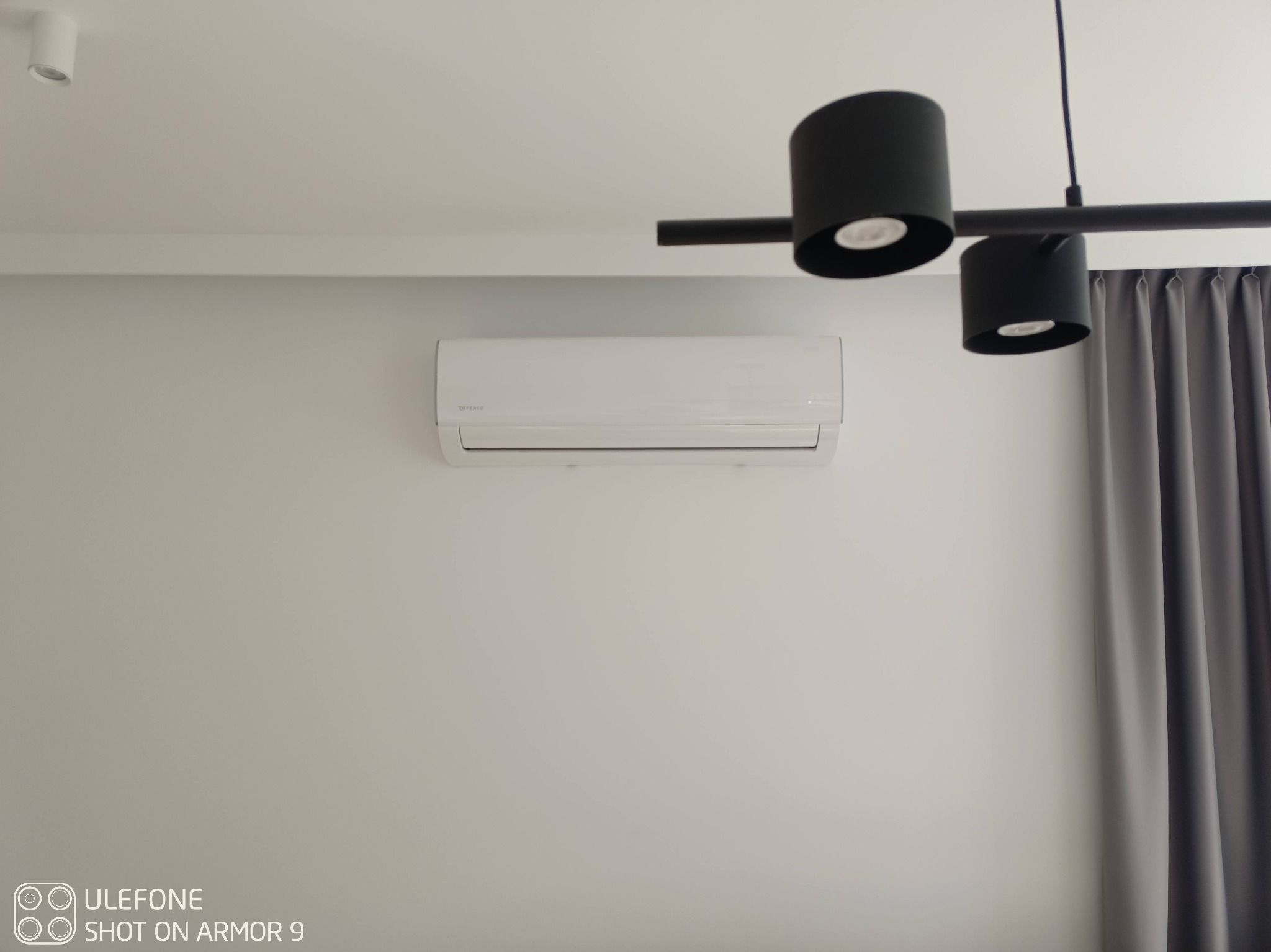 Biała jednostka wewnętrzna klimatyzatora zamontowana na jasnoszarej ścianie w nowoczesnym wnętrzu, z czarną lampą sufitową i szarymi zasłonami w tle.