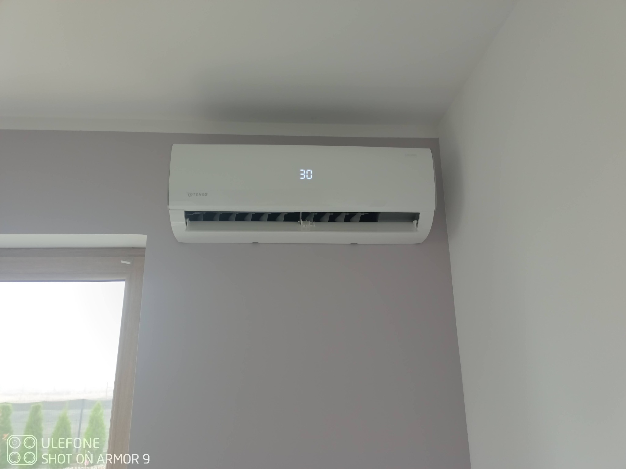Biały klimatyzator Rotenso z wyświetlaczem wskazującym temperaturę 30 stopni Celsjusza, zamontowany na szarej ścianie w pomieszczeniu z oknem.