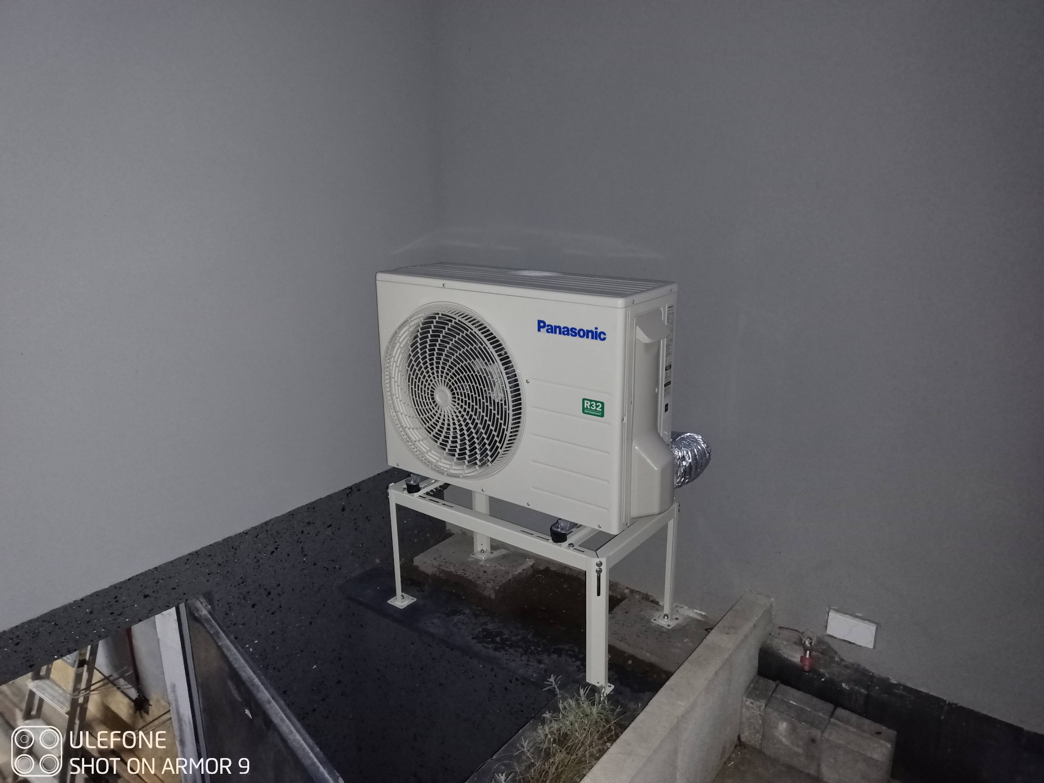 Zewnętrzna jednostka klimatyzacji Panasonic zamontowana na metalowym stelażu, z widocznym przewodem odprowadzającym ciepło i etykietą R32, na tle szarej ściany i fragmentu dachu.