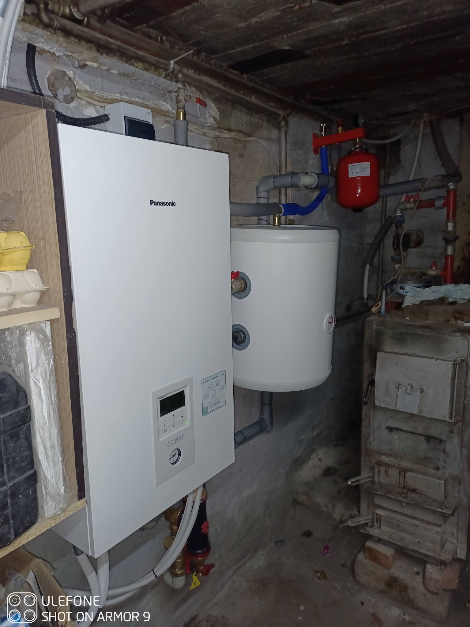 Nowa biała pompa ciepła Panasonic Aquarea zamontowana w piwnicy, obok starego pieca węglowego i bojlera. Widoczne rury i instalacje.