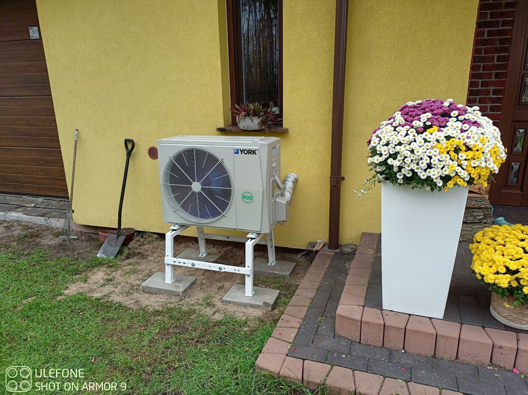 Zewnętrzna jednostka klimatyzacji York na białym stelażu, ustawiona na betonowych płytach obok żółtej ściany domu z kwiatami w donicach i narzędziami ogrodniczymi w tle.