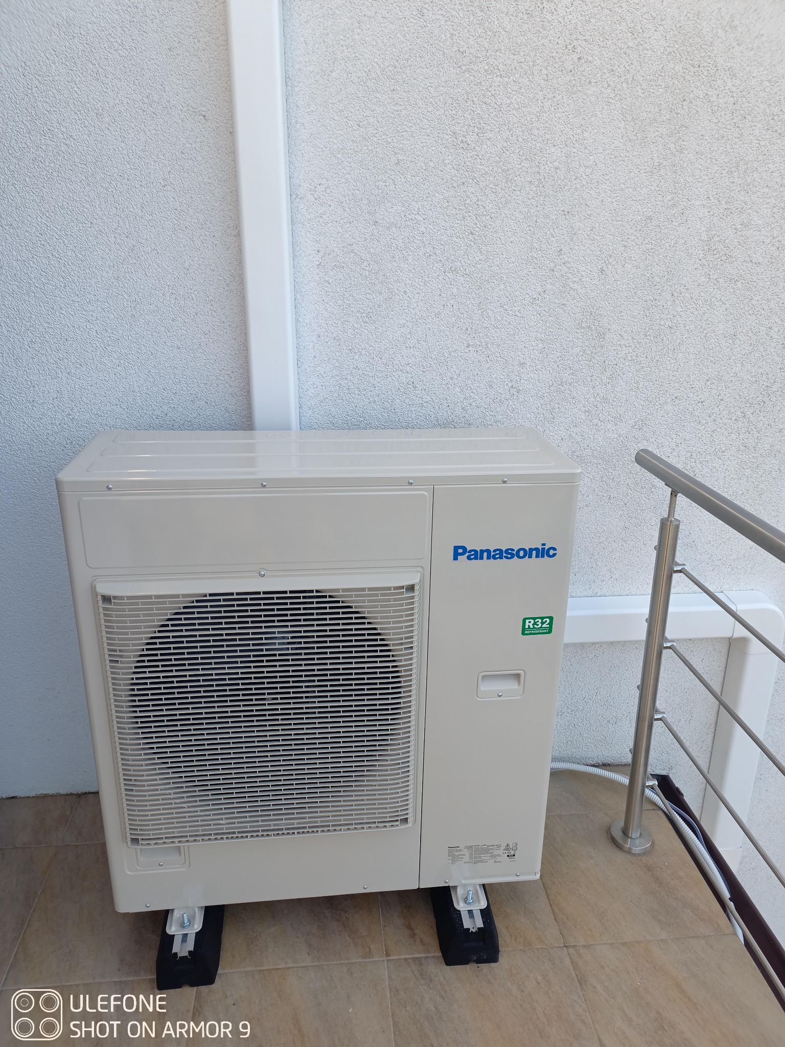 Zewnętrzna jednostka klimatyzacji Panasonic zamontowana na balkonie, widoczne przewody i mocowania, ściana z szorstkiego tynku.