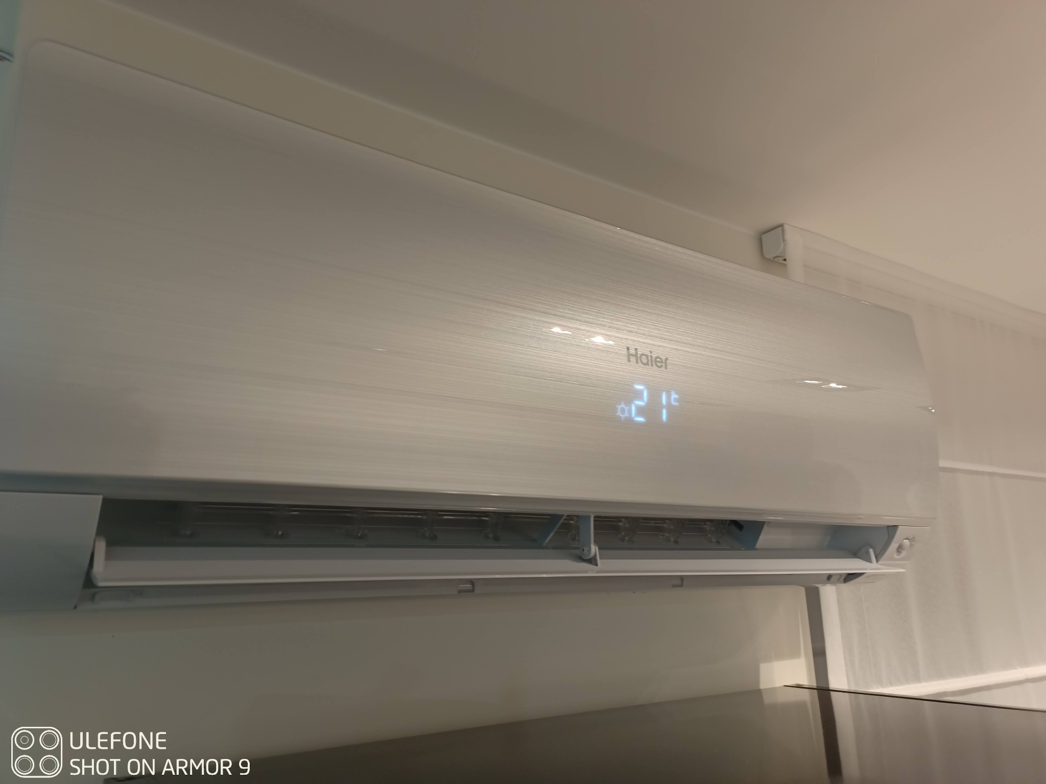 Biała jednostka wewnętrzna klimatyzatora Haier zamontowana na ścianie, wyświetlająca temperaturę 21 stopni Celsjusza.