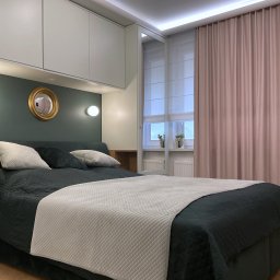 Pracownia Projektowa UNIQUE Anna Klimczewska-Waszczyk - Nowoczesna sypialnia z łóżkiem w odcieniach zieleni, białymi szafkami nad łóżkiem i okrągłym lustrem w złotej ramie. Parkiet w jodełkę i pudrowo-różowe zasłony.