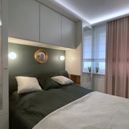 Pracownia Projektowa UNIQUE Anna Klimczewska-Waszczyk - Nowoczesna sypialnia w stonowanych barwach: oliwkowa ściana za łóżkiem, okrągłe lustro w złotej ramie, białe szafki pod sufitem i pudrowo-różowe zasłony.