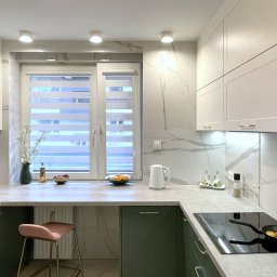 Pracownia Projektowa UNIQUE Anna Klimczewska-Waszczyk - Nowoczesna kuchnia z zielonymi szafkami, blatem imitującym marmur i oknem z roletami. Na blacie akcesoria kuchenne i dekoracje. Na płycie grzewczej patelnia z jedzeniem.