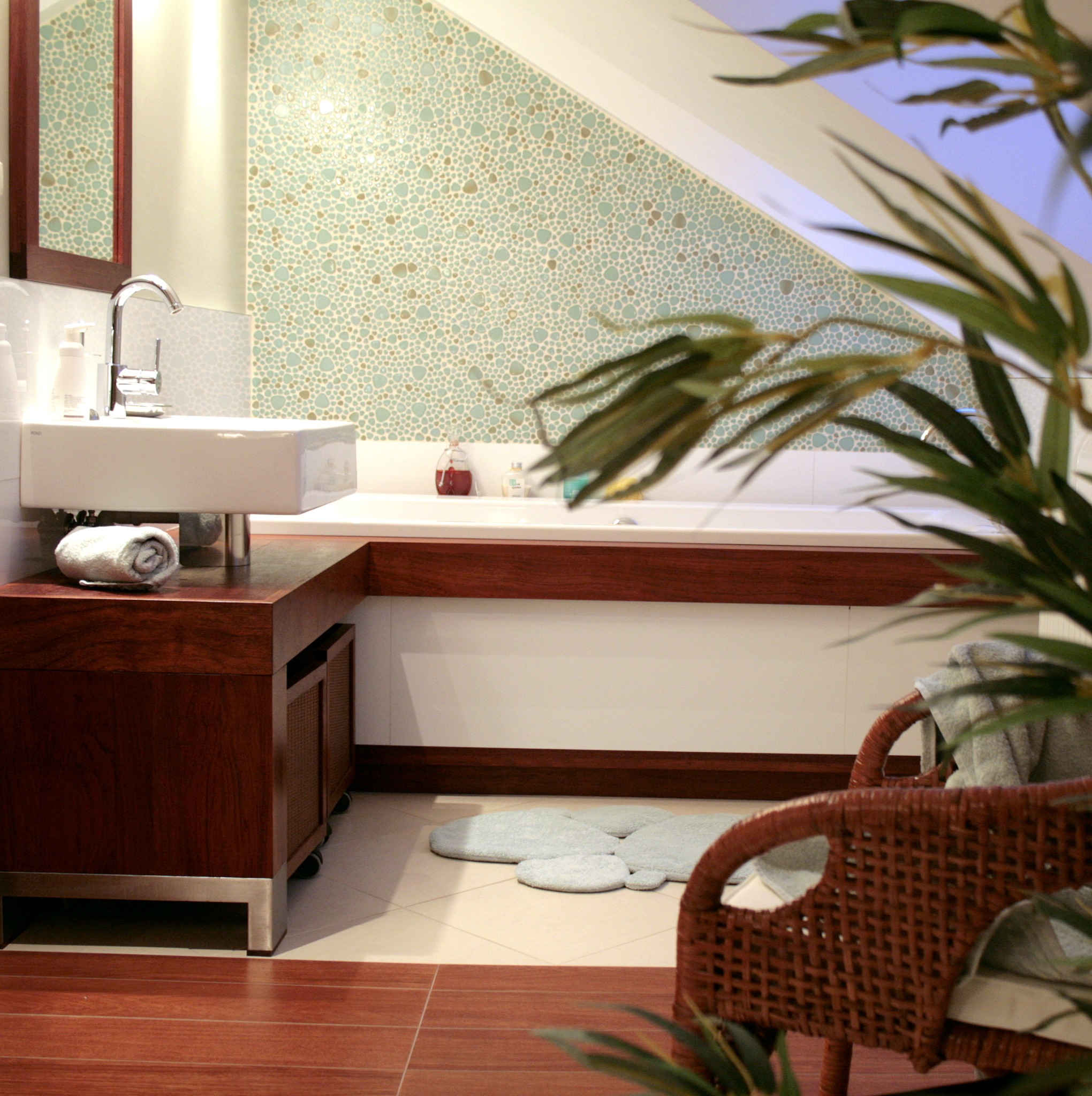 Jasna łazienka na poddaszu z wanną, umywalką nablatową i mozaikową ścianą, akcentowana rośliną doniczkową i wiklinowym fotelem.