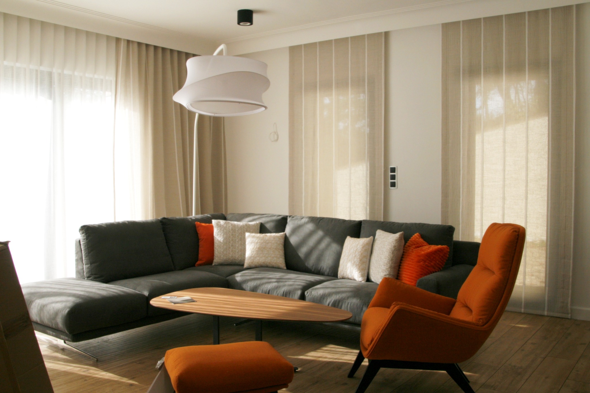 Nowoczesny salon z szarą sofą narożną, pomarańczowym fotelem i drewnianym stolikiem kawowym na tle jasnych zasłon i paneli okiennych.