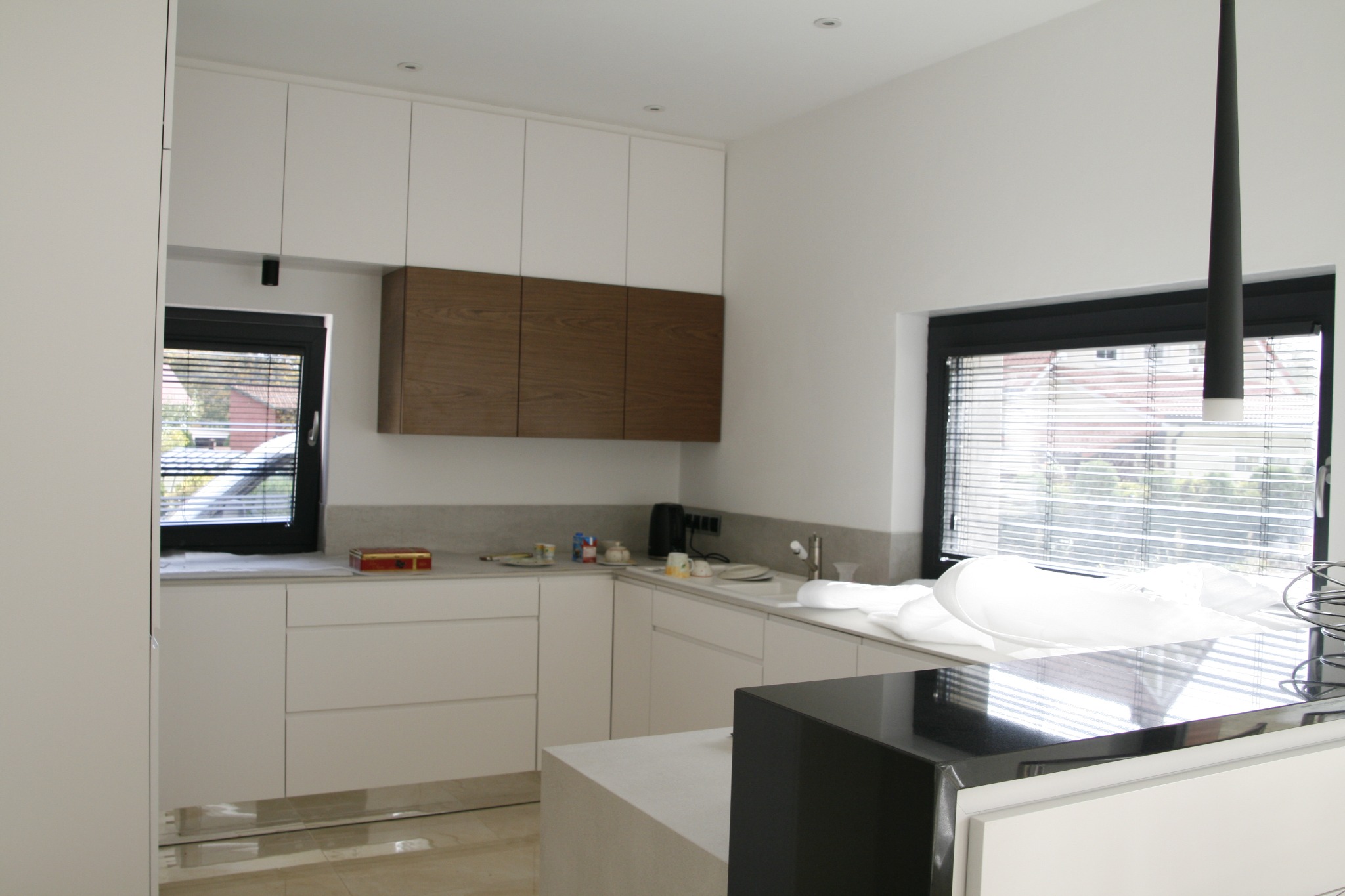 Nowoczesna kuchnia z białymi szafkami, drewnianymi akcentami i czarnymi ramami okiennymi, widziana z perspektywy wnętrza, z marmurową podłogą i minimalistyczną lampą wiszącą.