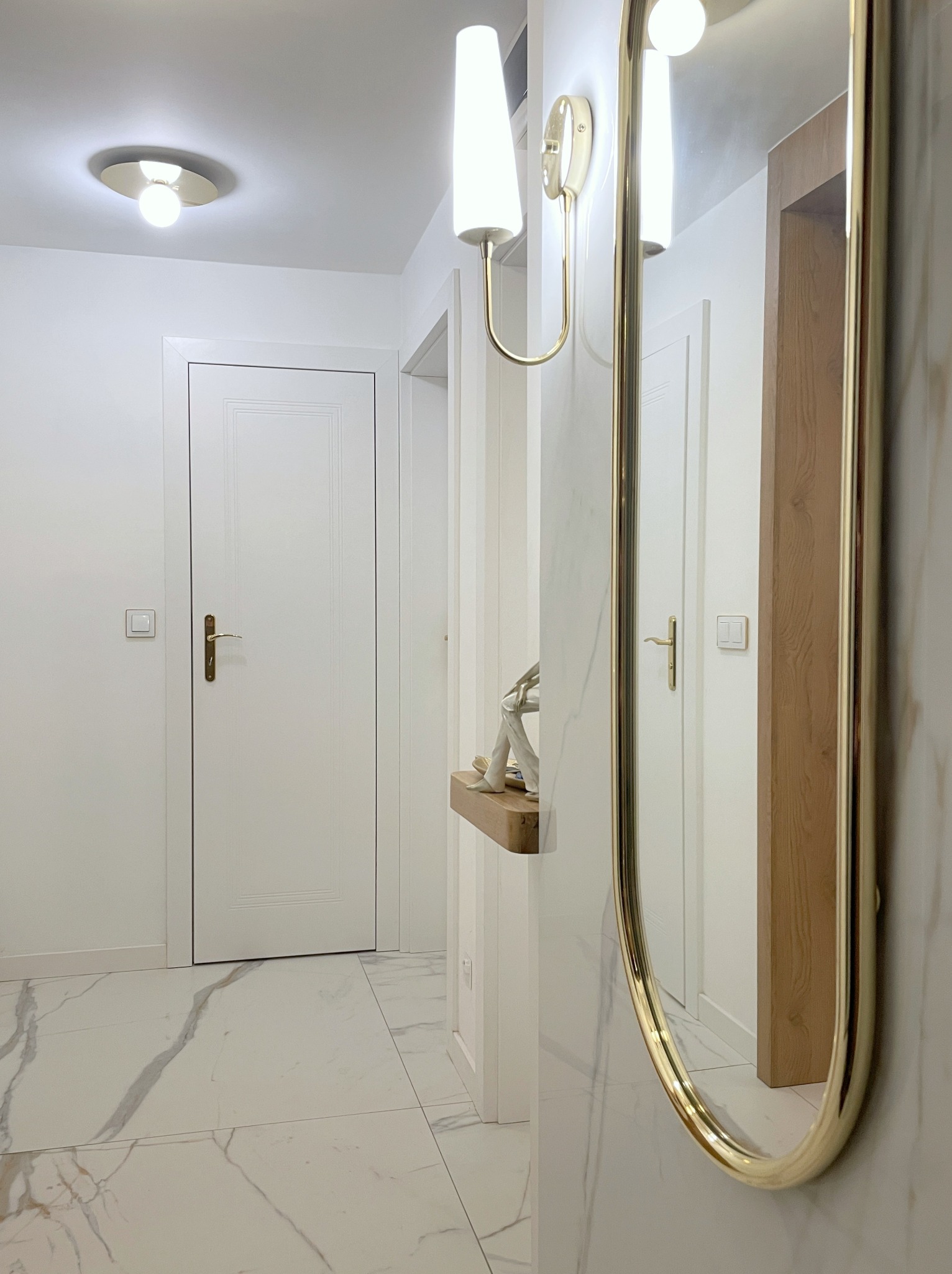 Nowoczesny przedpokój z marmurową podłogą, białymi drzwiami i złotym lustrem. Minimalistyczna konsola z dekoracją. Oświetlenie ścienne w stylu glamour.