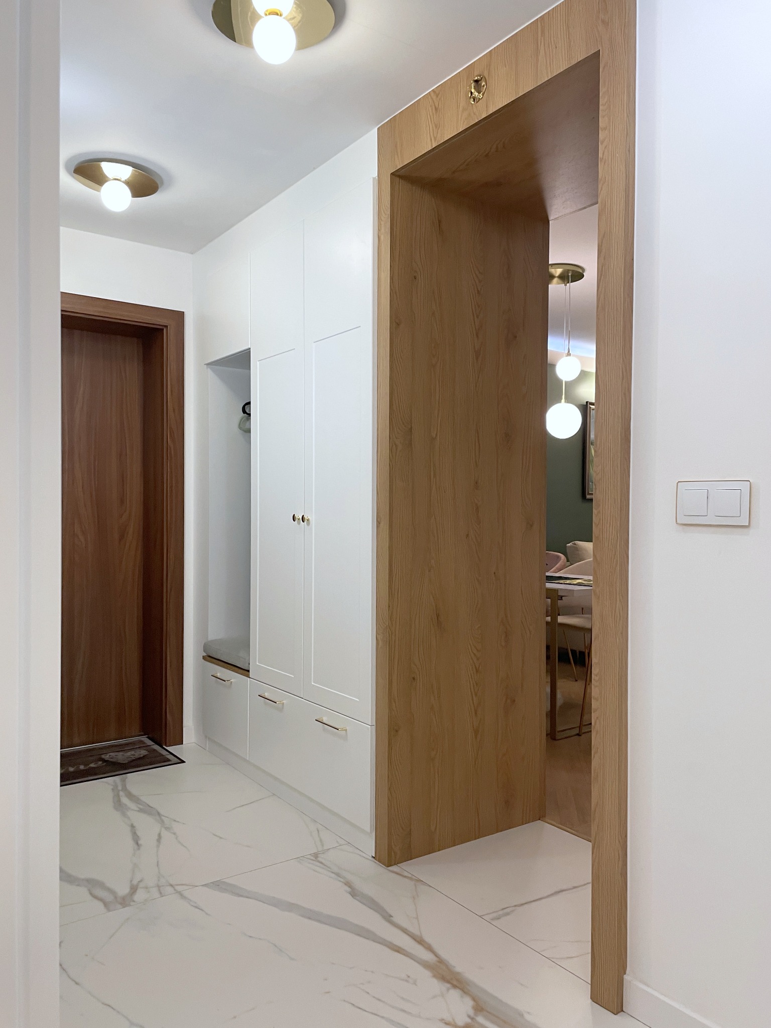 Nowoczesny przedpokój w Sopot z białą zabudową meblową, drewnianymi akcentami i marmurową podłogą. Widok na salon przez drewnianą framugę.