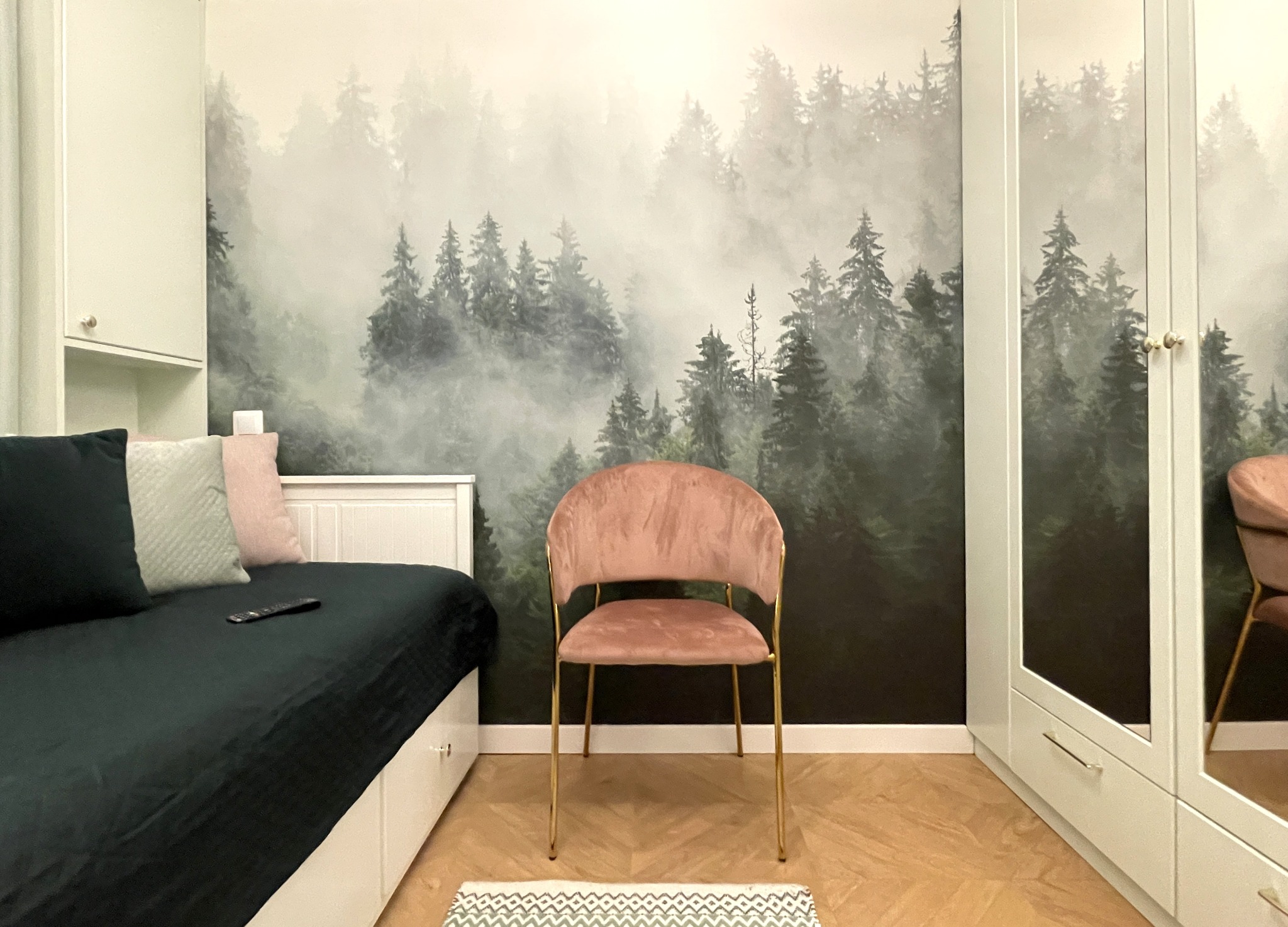 Pokój z łóżkiem, szafą i krzesłem na tle ściany z motywem leśnym. Jasne meble i drewniana podłoga tworzą przytulną aranżację wnętrza.