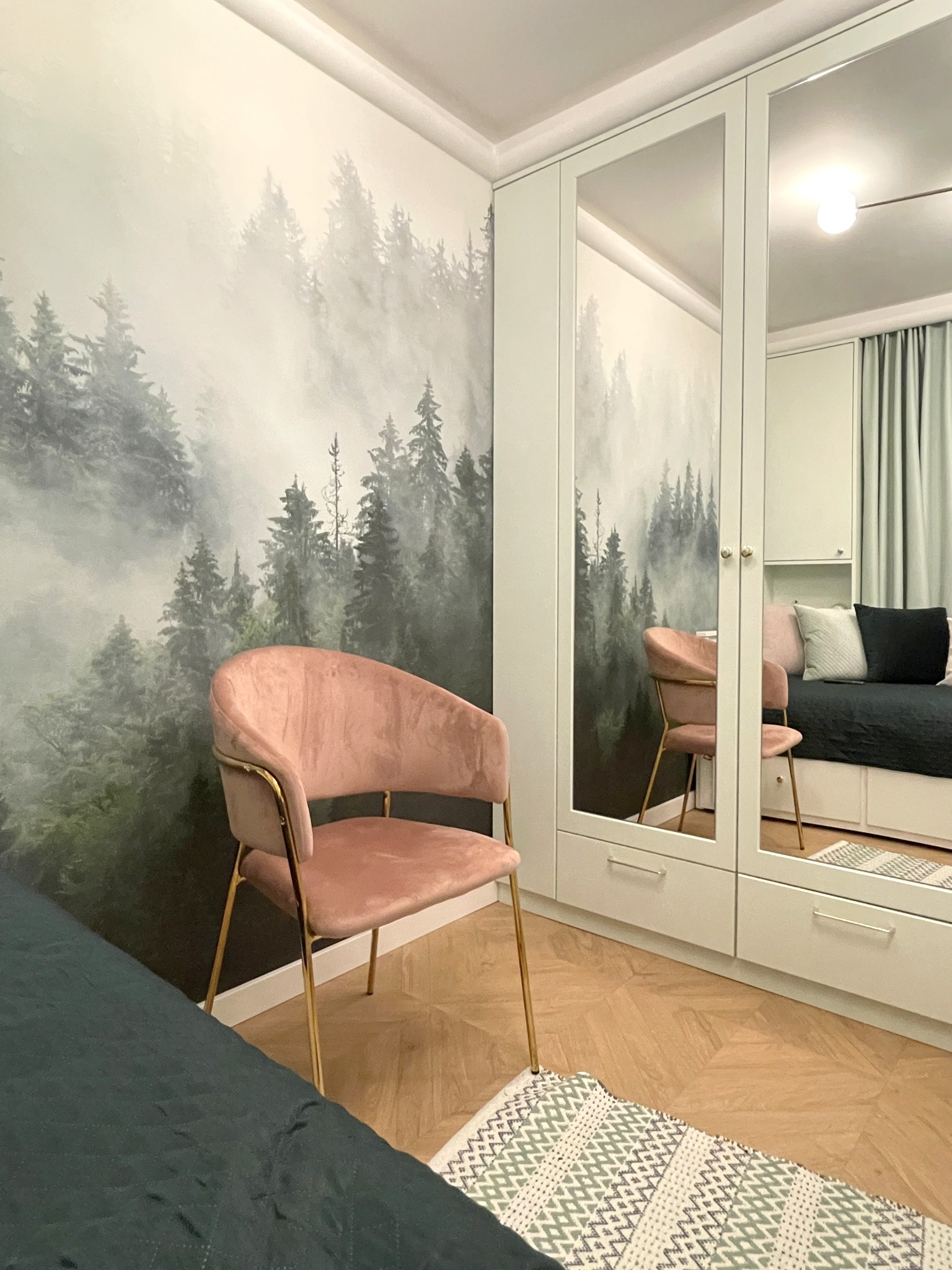 Pokój z fototapetą leśną, różowym krzesłem na złotych nogach i szafą z lustrami. Widoczny fragment łóżka i dywanik. Jasne, naturalne światło.