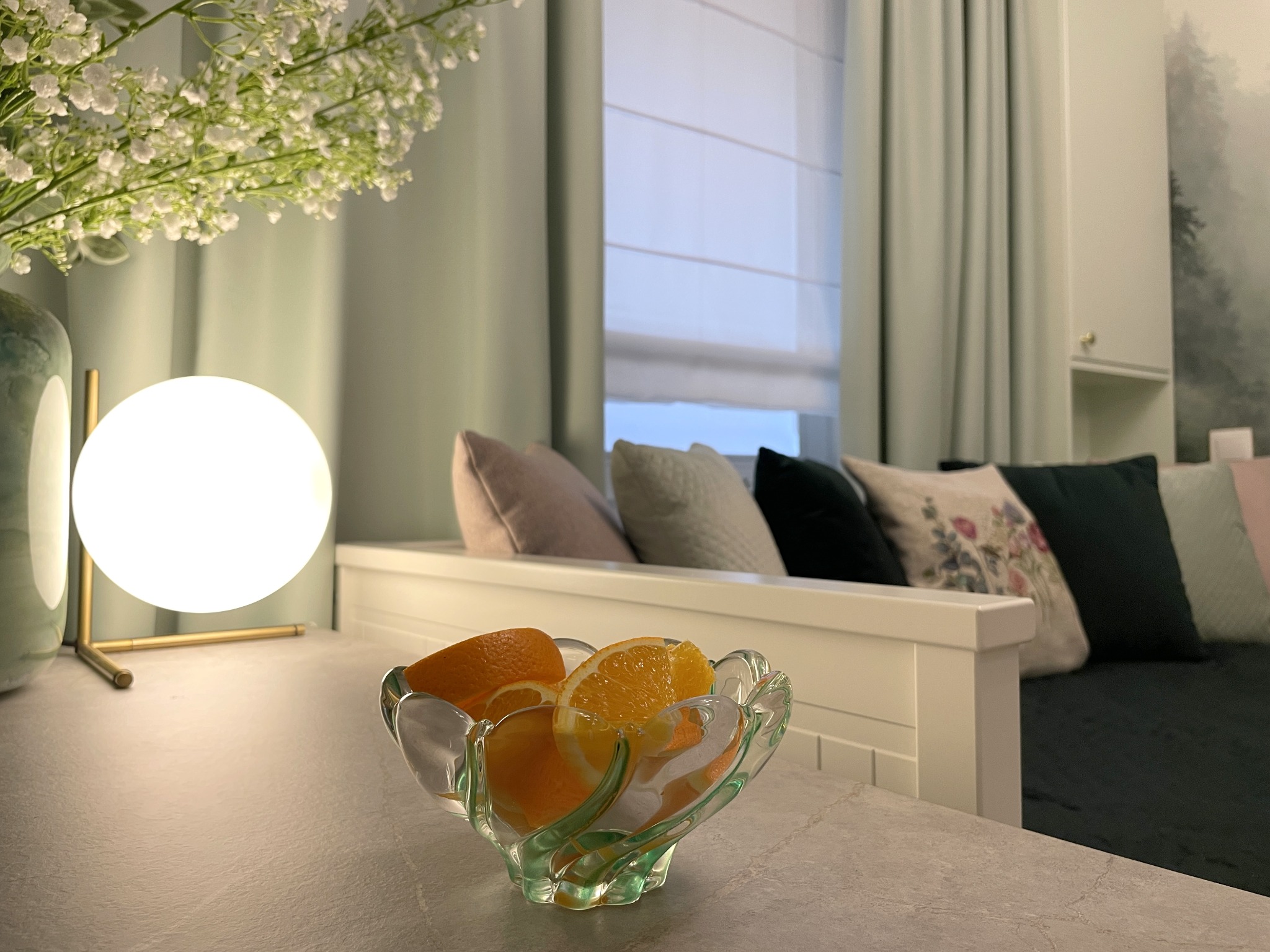 Stylowy pokój z dekoracyjną lampą kulą, kryształową misą z pomarańczami i kwiatami gipsówki. W tle sofa z poduszkami i zasłony w odcieniach zieleni.